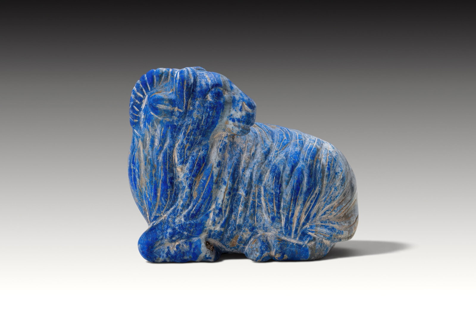 RARE MESOPOTAMIAN LAPIS LAZULI RAM FIGURINE: Ca. 2200 - 1800 BC. A lapis lazuli figurine carved as a recumbent ram with the legs tucked beneath the body. The head is turned to the side, with rounded eyes, a short muzzle, and ridged horns curving