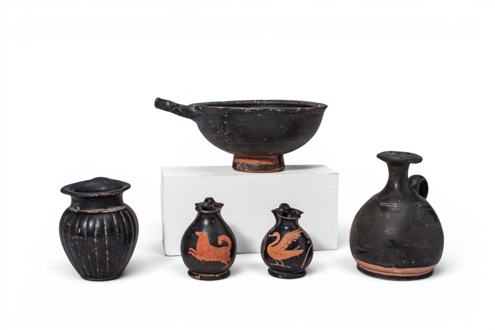 COLLECTION OF GREEK BLACK-GLAZED AND RED-FIGURE VESSELS: Ca. 400 BC. A collection of five black-glazed and red-figure vessels, including, from left to right: a cup with a deep rounded bowl, short foot and single horizontal handle; a ribbed mug with a rounde