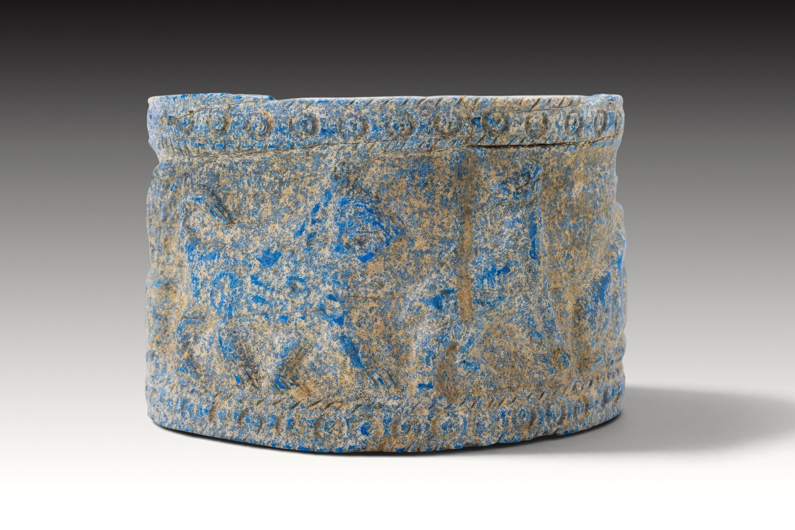 MESOPOTAMIAN LAPIS LAZULI PYXIS WITH ANIMALS AND TREES: Ca. 2200 - 1800 BC. A lapis lazuli pyxis with a cylindrical body, straight walls, and a flat circular base. The exterior is carved in low relief with animals standing among stylised trees, framed by n