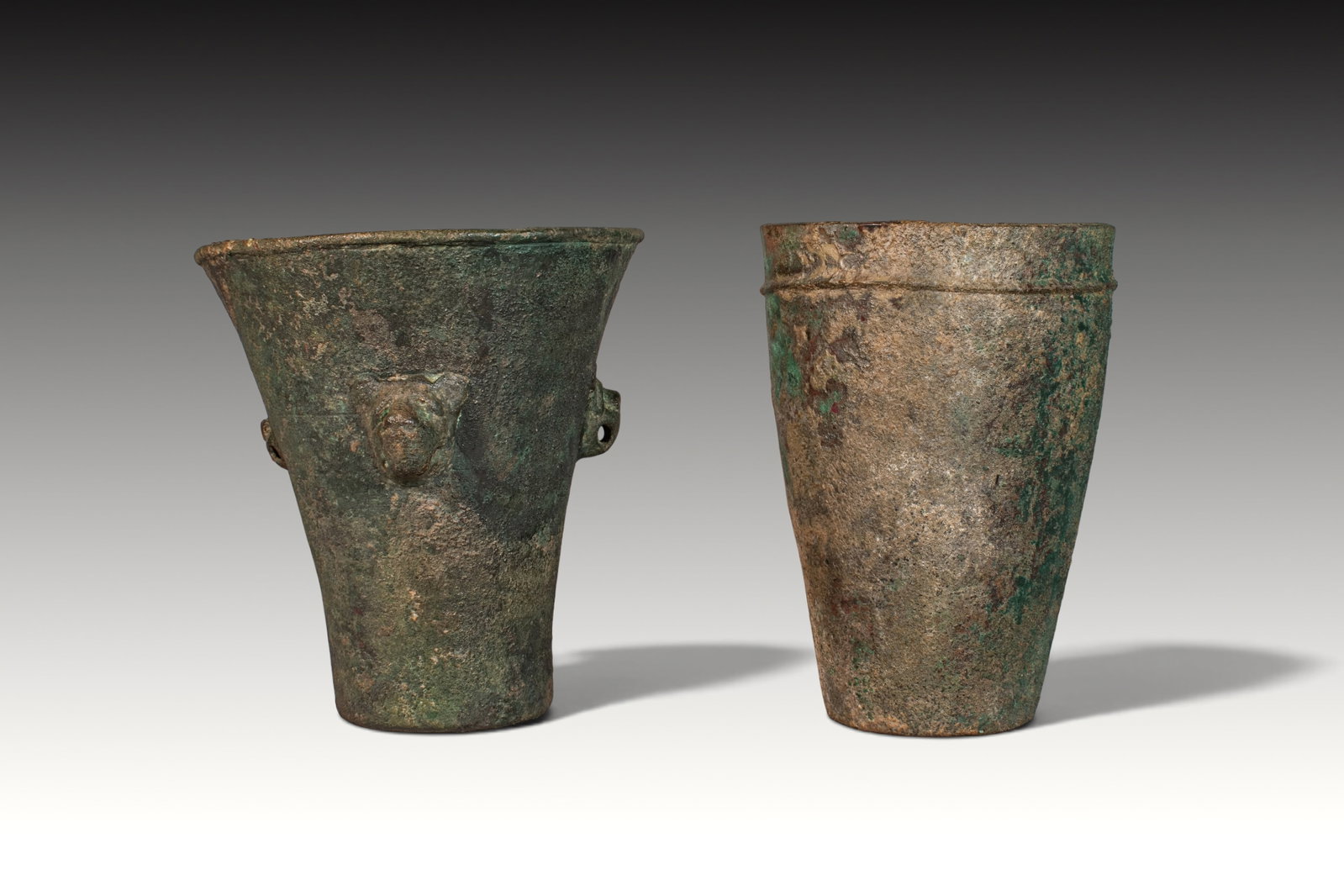 PAIR OF TALL BRONZE URARTIAN BEAKERS: Ca. 800 BC. A pair of tall bronze beakers, each with a tapering cylindrical body, flat base, and broad open mouth. The beaker on the left has a flaring rim and applied lion-head protomes set on the sh