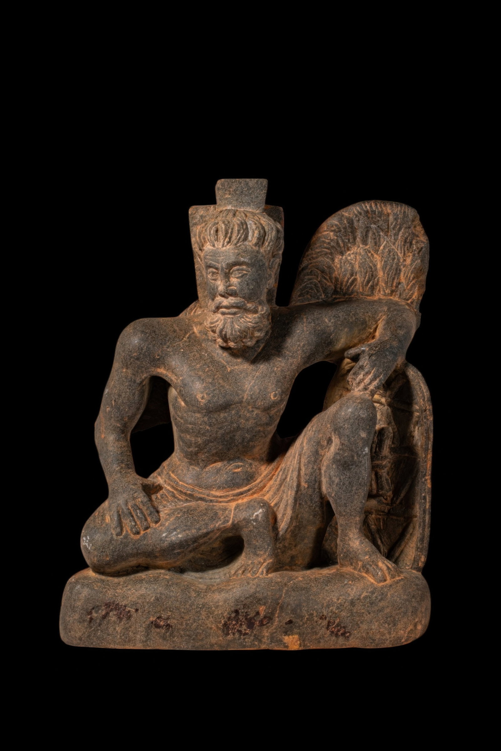 GANDHARAN STONE FIGURE OF ATLAS: Ca. 2nd - 4th century AD. A carved figure depicting the muscular figure of Atlas seated in a dynamic pose, with one leg folded beneath him and the other raised. The bearded figure is shown bare-cheste