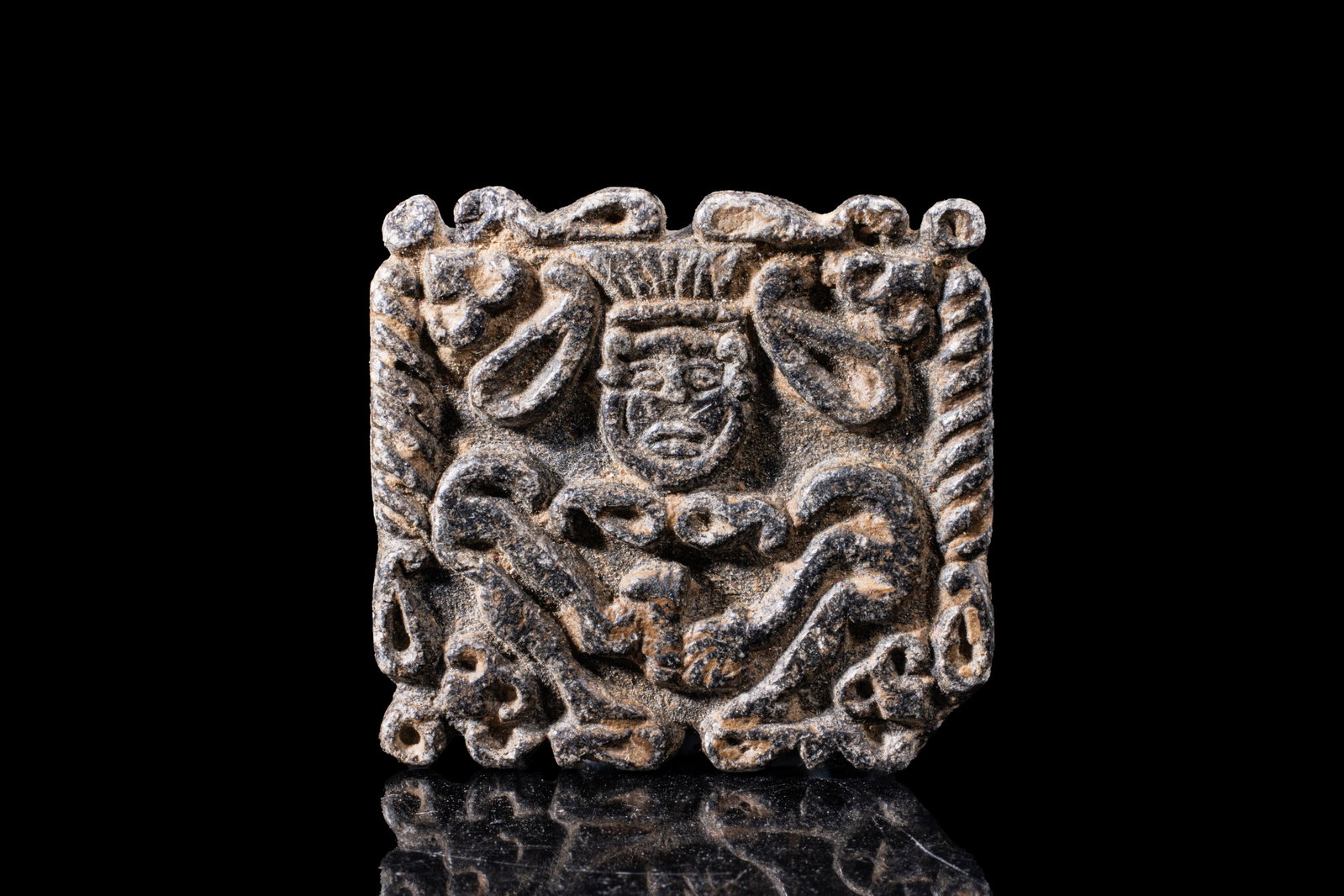 ROMANO-EGYPTIAN STONE PLAQUE OF BES: Ca. 30 BC - AD 200. A rectangular stone plaque carved with Bes wearing a tall feathered headdress and squatting frontally. The surrounding field is filled with ornamental motifs and enclosed by a twis