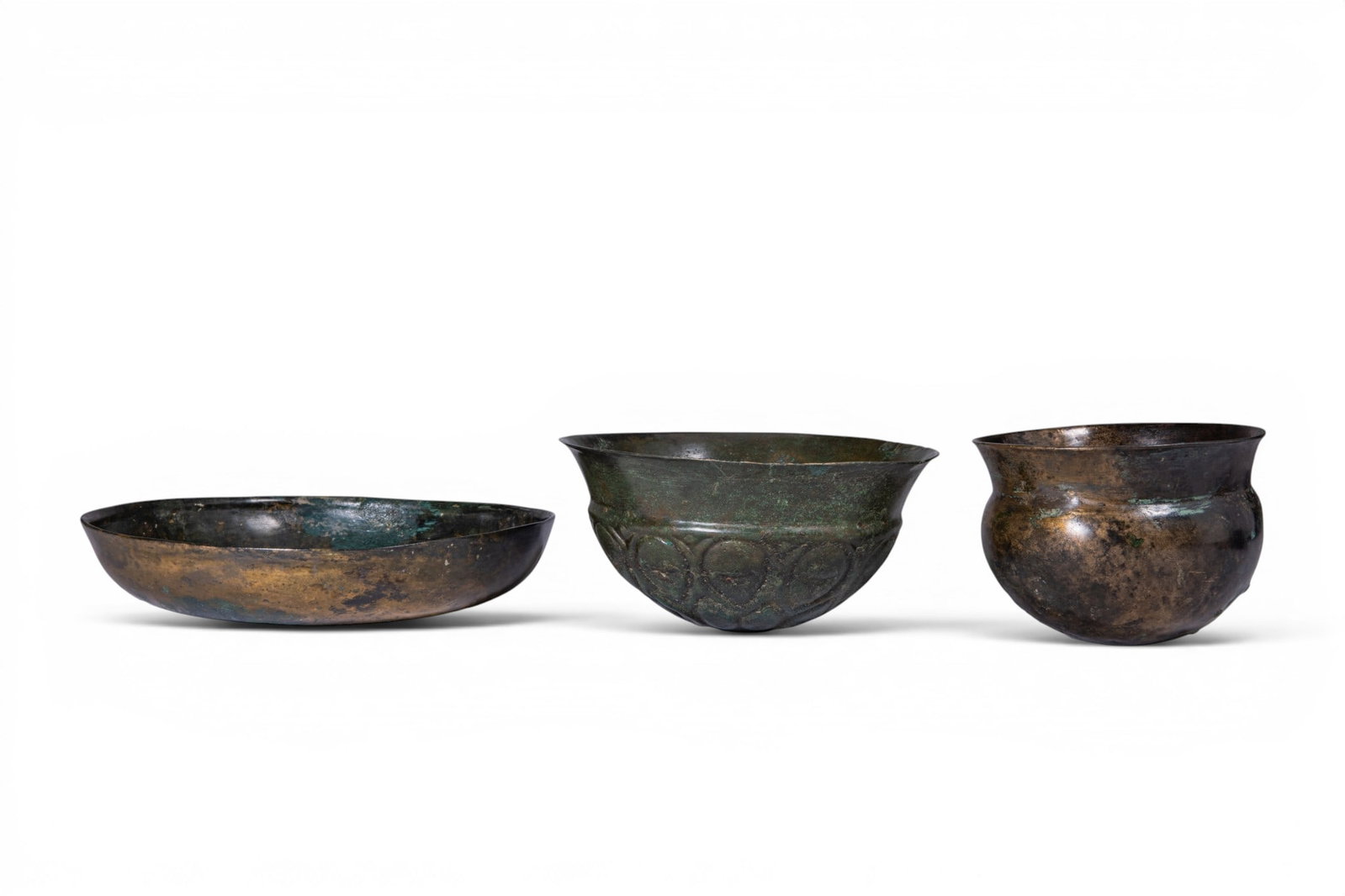 COLLECTION OF GREEK BRONZE BOWLS: Ca. 500 - 300 BC. A collection of three bronze vessels, including, from left to right: a shallow bowl with rounded sides and an omphalos; a bowl with a flaring mouth and a lower body decorated with pe