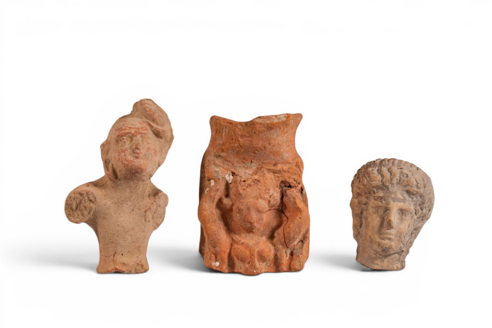 COLLECTION OF GREEK TERRACOTTA RITUAL STATUETTES: Ca. 400 - 200 BC. A collection of three terracotta ritual statuettes, including, from left to right: a male torso with a rounded face beneath a side cap; a rectangular plaque depicting a female bust w