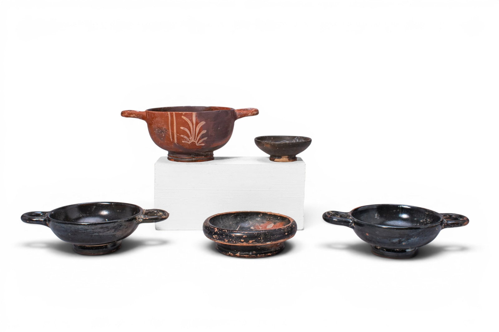 COLLECTION OF GREEK TERRACOTTA VESSELS: Ca. 400 - 300 BC. A collection of five terracotta vessels, including, from left to right: a black-glazed kylix with a shallow bowl, twin horizontal handles, and low foot, decorated with palmettes on