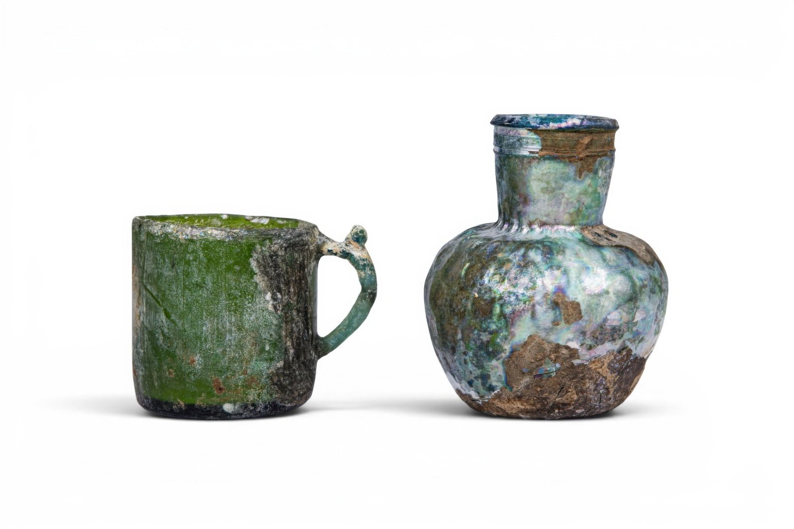 PAIR OF UMAYYAD GLASS VESSELS: Ca. AD 800 - 950. A pair of glass vessels, comprising, from left to right: a green glass mug with straight sides and a loop handle terminating in a thumb rest; and a pale green glass bottle with a glo