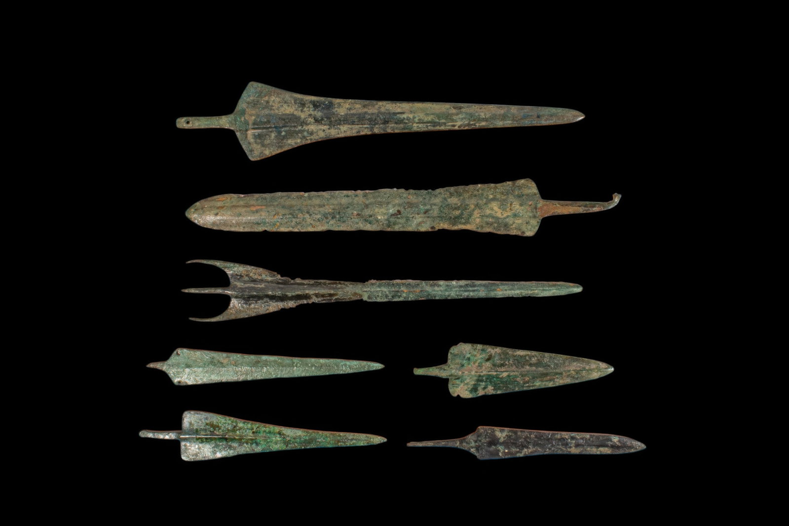 LARGE COLLECTION OF BRONZE AGE / EARLY GREEK PERIOD BRONZE DAGGERS AND SPEARHEADS: Ca. 1200 - 800 BC. A collection of seven bronze daggers and spearheads, including, from top to bottom: a spearhead with a long tapering blade, flaring shoulders, midrib, and short tang; a broad dagger