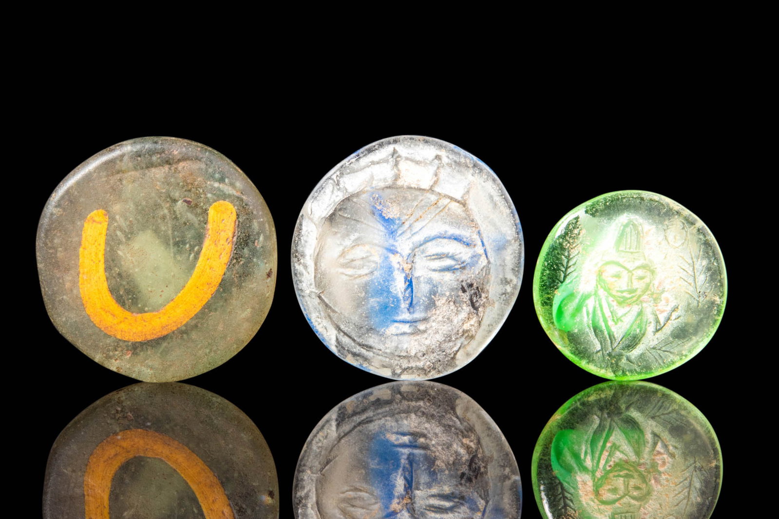 RARE ROMANO - EGYPTIAN GLASS STAMPED DISC AMULETS: Ca. 30 BC - AD 200. A set of rare Romano-Egyptian glass stamped disc amulets. The first shows a U-shaped sign in gold; the second depicts a frontal face of a man with a hood; the third shows a bust of