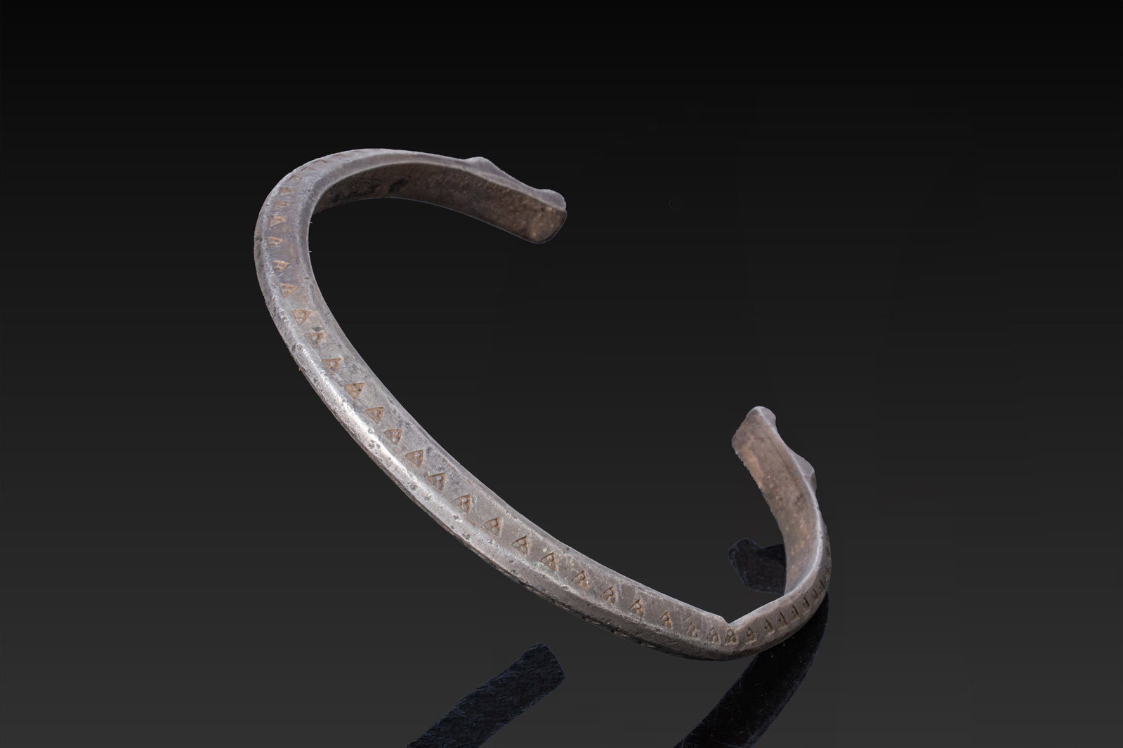 STUNNING VIKING SILVER BRACELET WITH FAFNIR HEAD FINIALS: Ca. AD 700 - 1100. A silver penannular bracelet with a carinated hoop tapering to stylised Fafnir head finials. The outer face is decorated with two rows of stamped triangular motifs with three dots i
