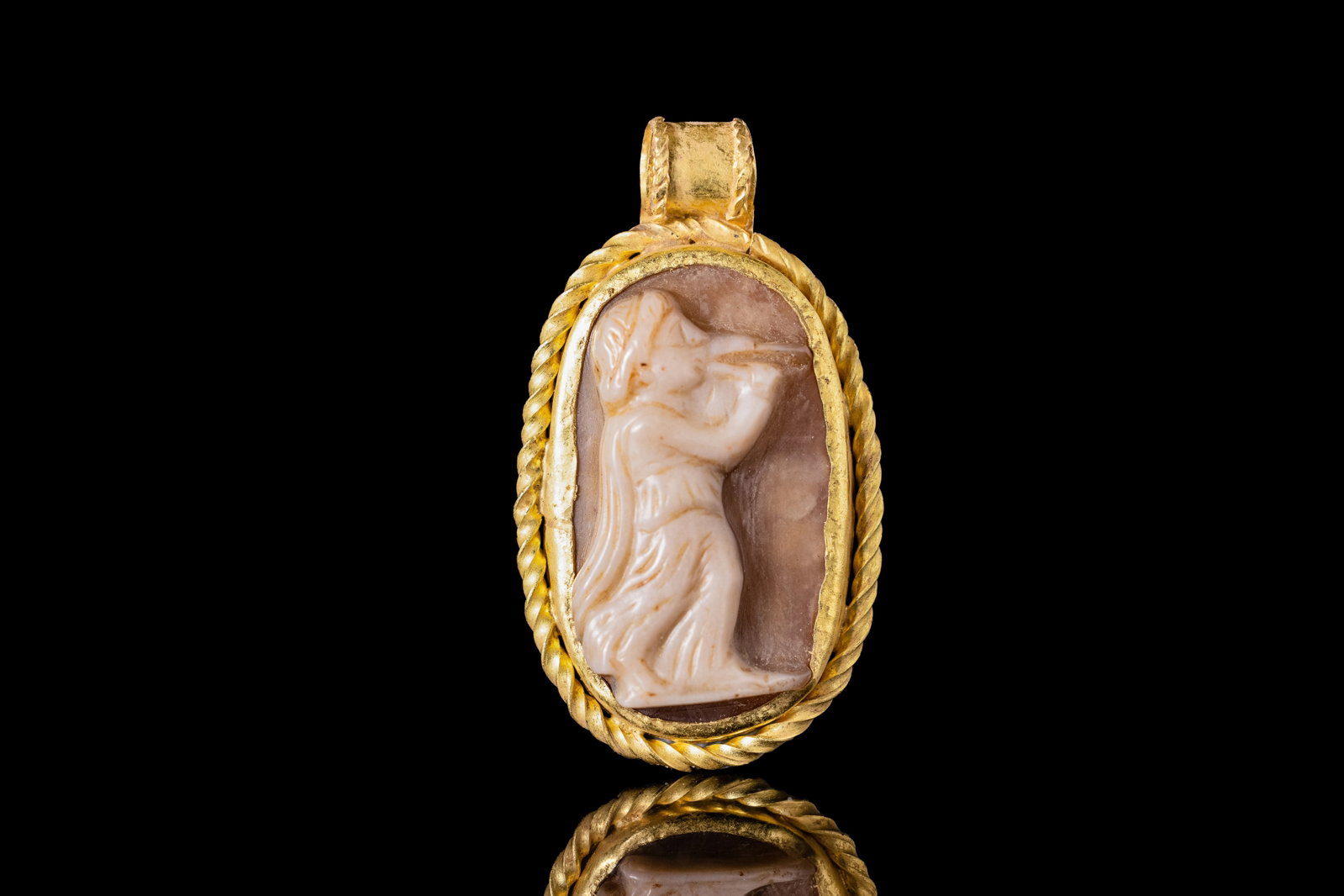 ROMAN AGATE CAMEO OF A MUSICIAN PLAYING FLUTE IN GOLD PENDANT: Ca. AD 1 - 300. An agate cameo carved with a draped musician in profile, the head inclined towards a double flute held to the mouth. The figure is framed within a possibly later gold mount with a plai