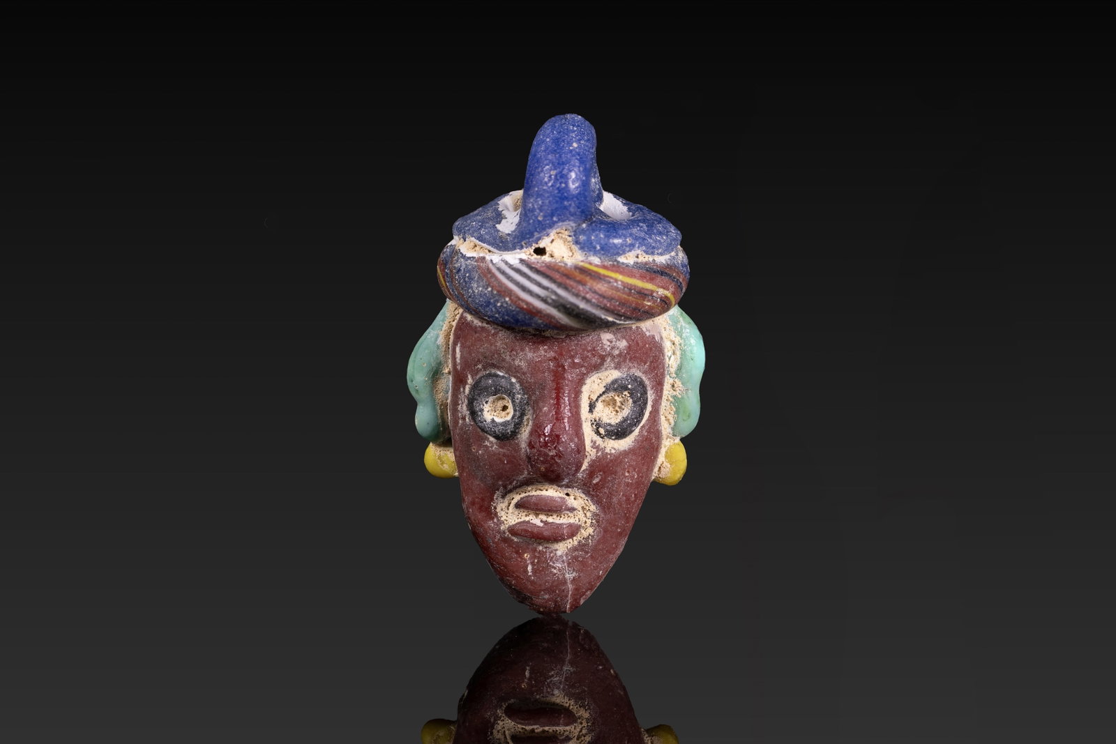 PHOENICIAN POLYCHROME GLASS FACE PENDANT: Ca. 600 - 300 BC. A glass face pendant with a deep red face, black ringed eyes, an elongated nose and a small mouth. Turquoise ears are set with yellow earrings, and a broad blue headdress is edged be