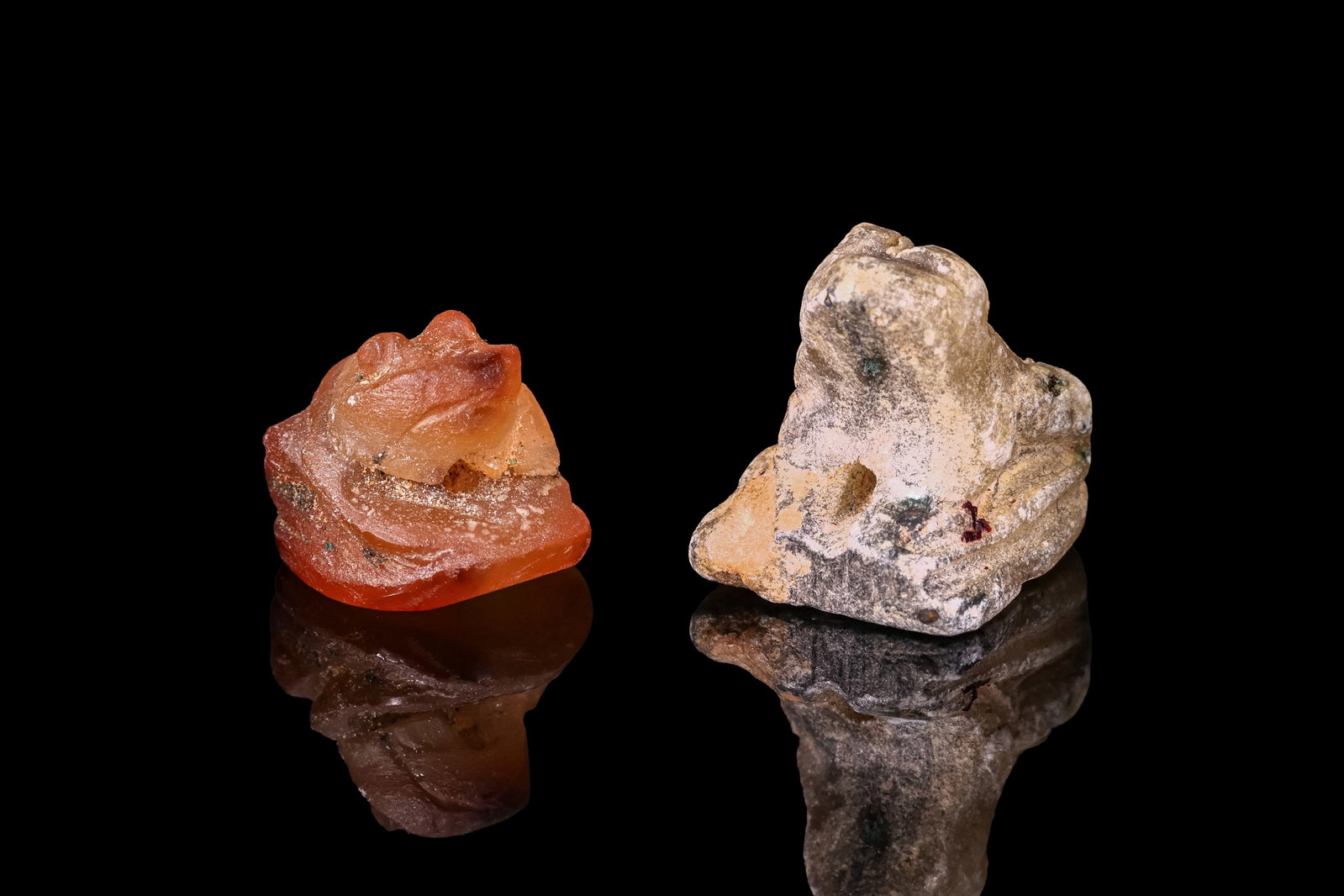 PAIR OF EGYPTIAN STONE FROG AMULETS: Late Period, Ca. 664 - 332 BC. A pair of stone frog amulets, each carved crouching with the legs folded against the body and perforated through the base. The one on the left is in carnelian, with roun