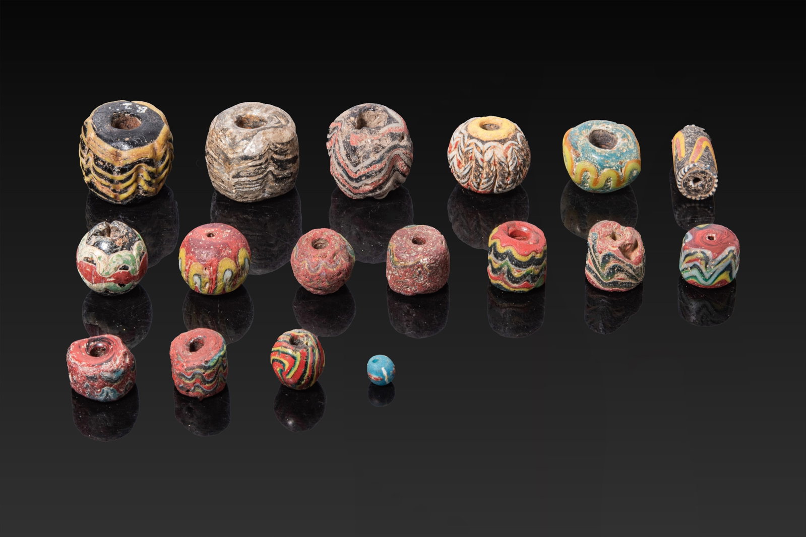 COLLECTION OF ROMANO-EGYPTIAN POLYCHROME GLASS BEADS: Ca. 200 BC - AD 200. A collection of 17 polychrome glass beads, comprising rounded, barrel and cylindrical types in varied sizes. The group includes wound glass beads with trailed and combed decoratio