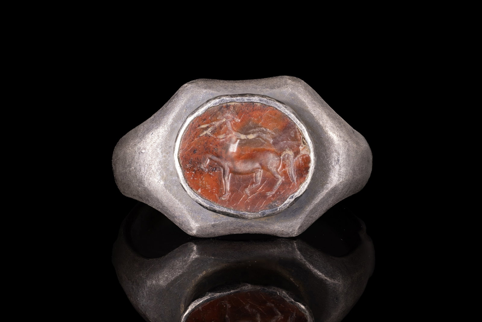ROMAN CARNELIAN INTAGLIO WITH CENTAUR PLAYING FLUTE IN SILVER RING: Ca. AD 300 . A carnelian intaglio engraved with a centaur moving in profile, its right foreleg raised and a flute held to its mouth. The intaglio is set in a possibly later silver ring with a slender 