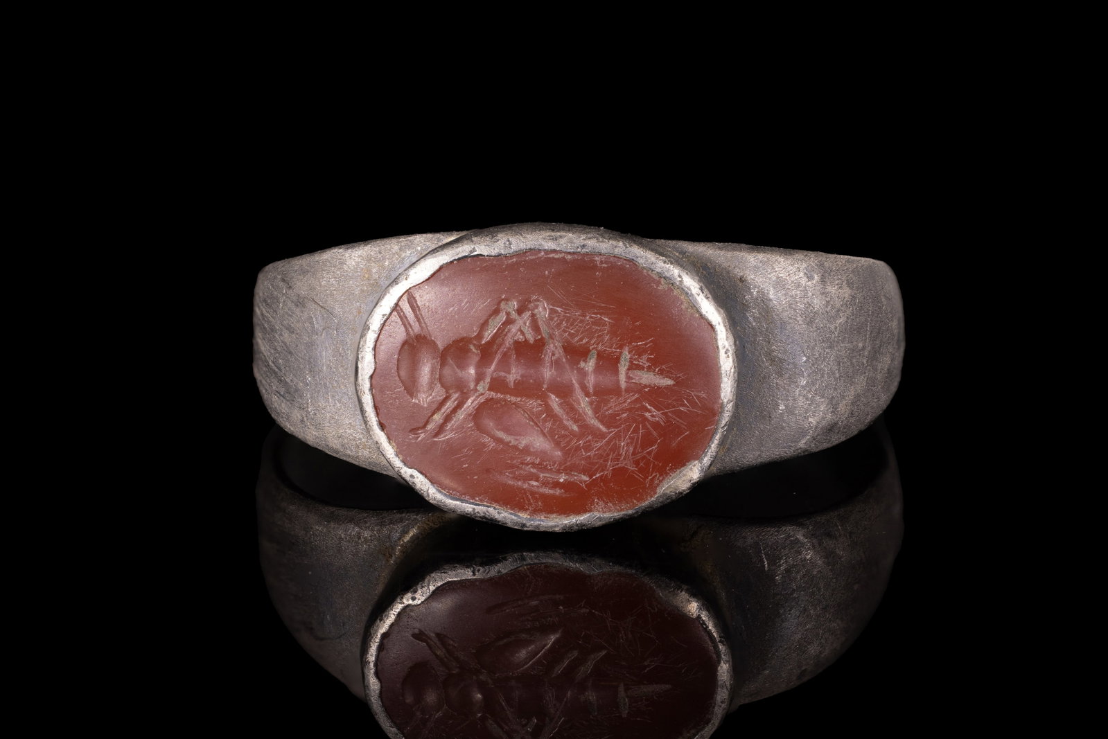 ROMAN CARNELIAN INTAGLIO WITH GRASSHOPPER IN SILVER RING: Ca. AD 1 - 300. A carnelian intaglio engraved with a grasshopper in profile, with long antennae, folded legs and a segmented body. The intaglio is set in a possibly later silver ring with a flat-secti