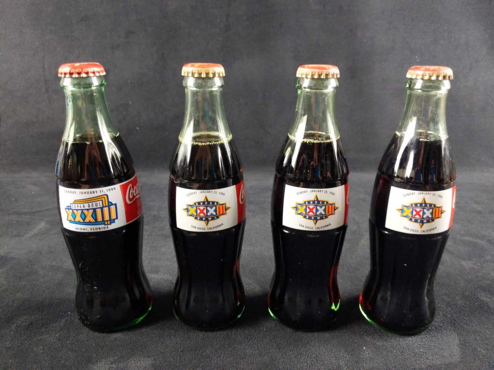 Four Full Coca Cola Bottles Superbowl: This lot consists of: (3) Superbowl XXXII - 1998. (1) Superbowl XXXIII - 1999. Bottles are approx 2" x 2" x 7 1/2". Bottles are in good, used condition with some wear on the bottle caps. Please see im