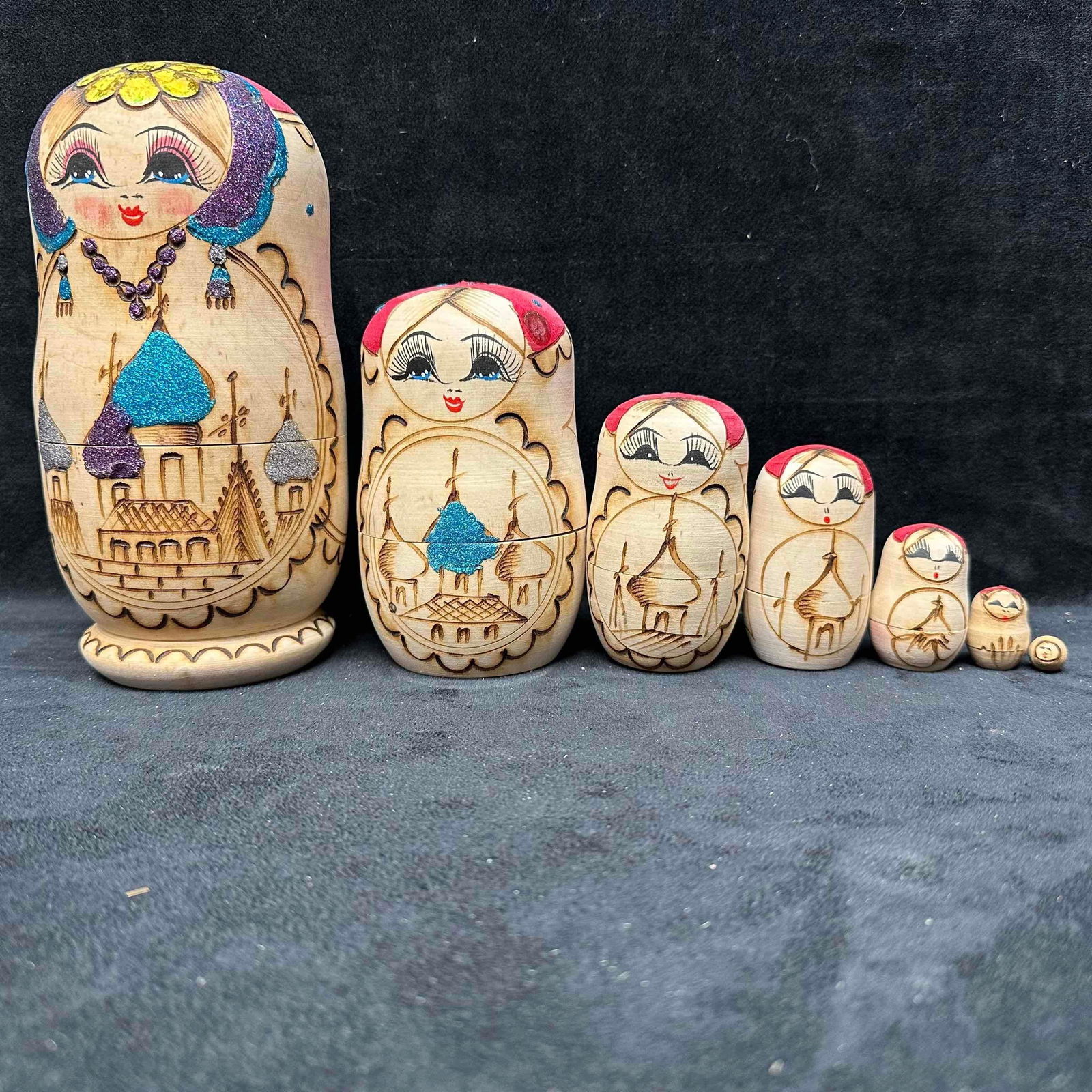 Russian Matryoshka Nesting Dolls Seven Piece Hand Crafted Set: Set of 7 traditional Russian nesting dolls, hand-painted with etched floral and architectural designs accented in glitter. The largest doll depicts a woman in a headscarf with cathedral scenes, while