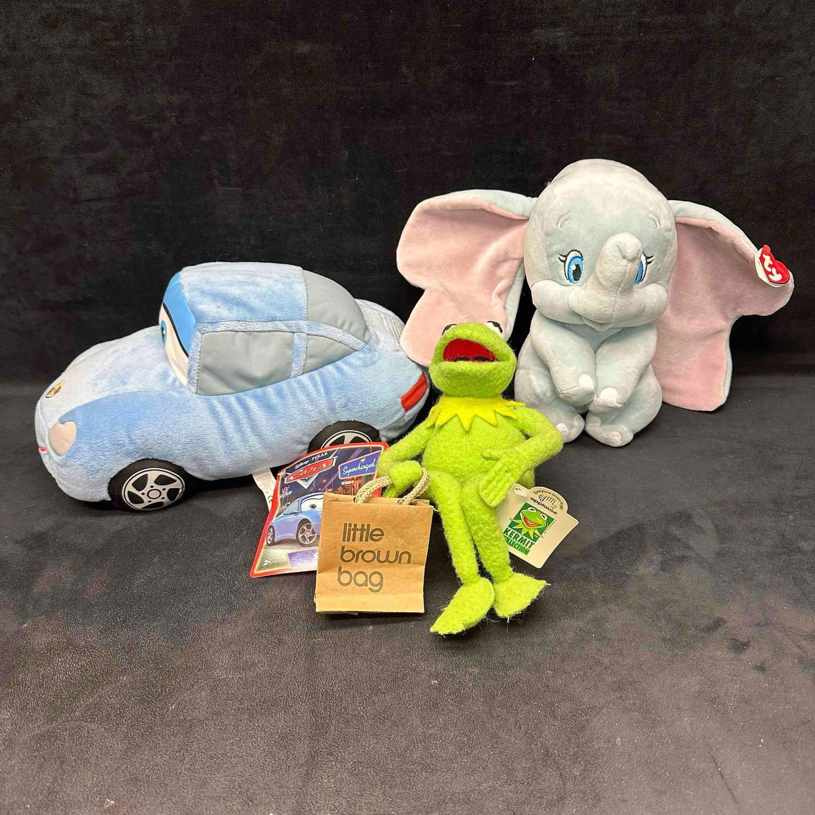 Three 3 Plush Toys Disney Cars Sally Ty Dumbo and Applause Kermit with Brown Bag Set: Group of three Disney and Jim Henson character plush toys including: Disney/Pixar Cars “Sally” plush with tag Ty Beanie Baby “Dumbo” plush with tag Applause Kermit plush with original