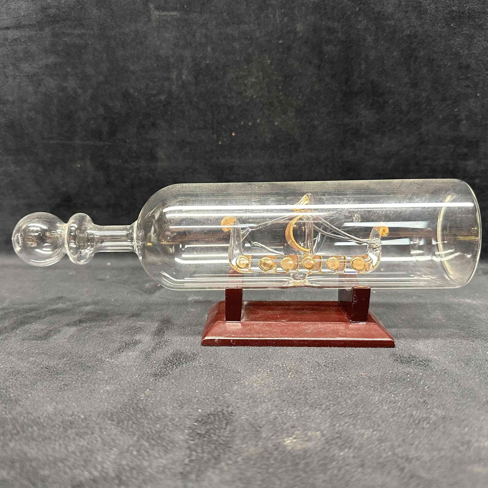 Intricate Art Glass Ship in a Bottle with Wood Stand: Clear glass bottle containing a finely crafted glass ship with gold-toned details, displayed on a wooden stand. The ship features a curved hull, round shields, and a single mast with sail, evoking a V