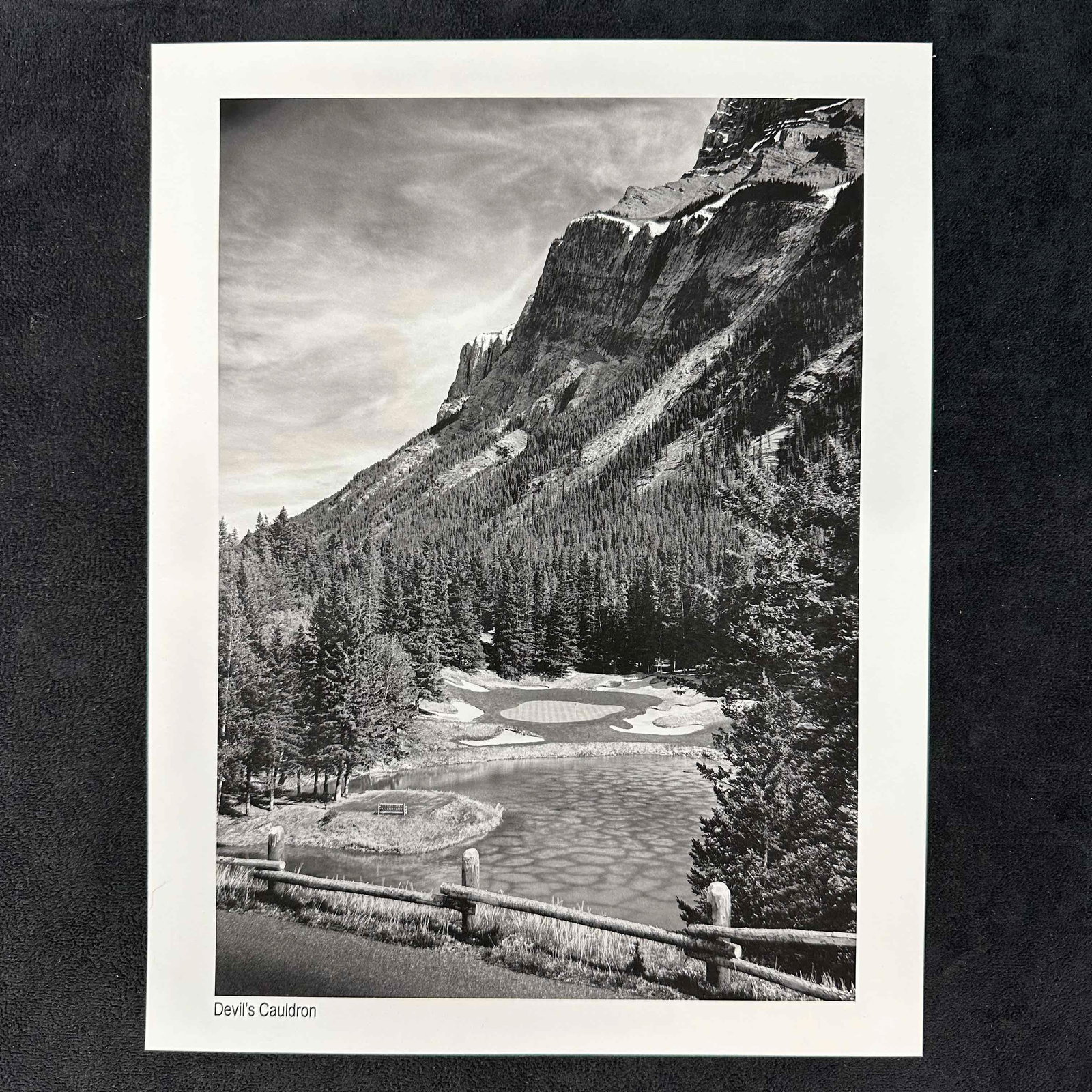 Devil's Cauldron at Banff Springs Golf Course Black & White Photography Print: Black-and-white photographic print titled "Devil’s Cauldron." Features a dramatic golf course scene nestled at the base of a towering mountain with dense forest and a reflective water hazard