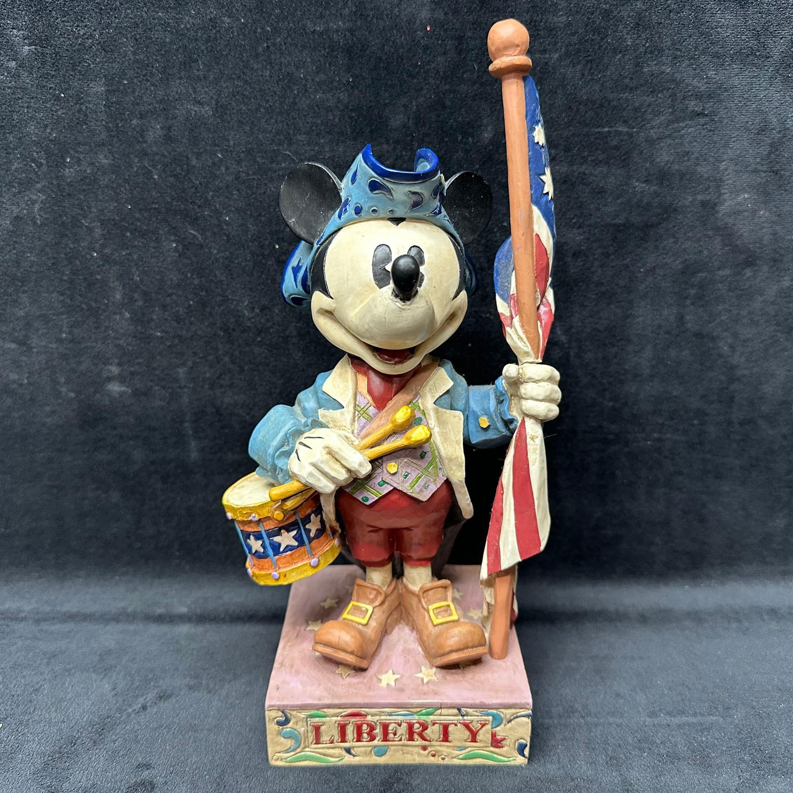 Disney Traditions by Jim Shore The Ultimate Patriot Mickey Mouse Figurine: A 2005 Disney Traditions by Jim Shore The Ultimate Patriot figurine featuring Mickey Mouse dressed in colonial attire, holding a patriotic flag with a drum at his side. Part of the Walt Disney Showcas