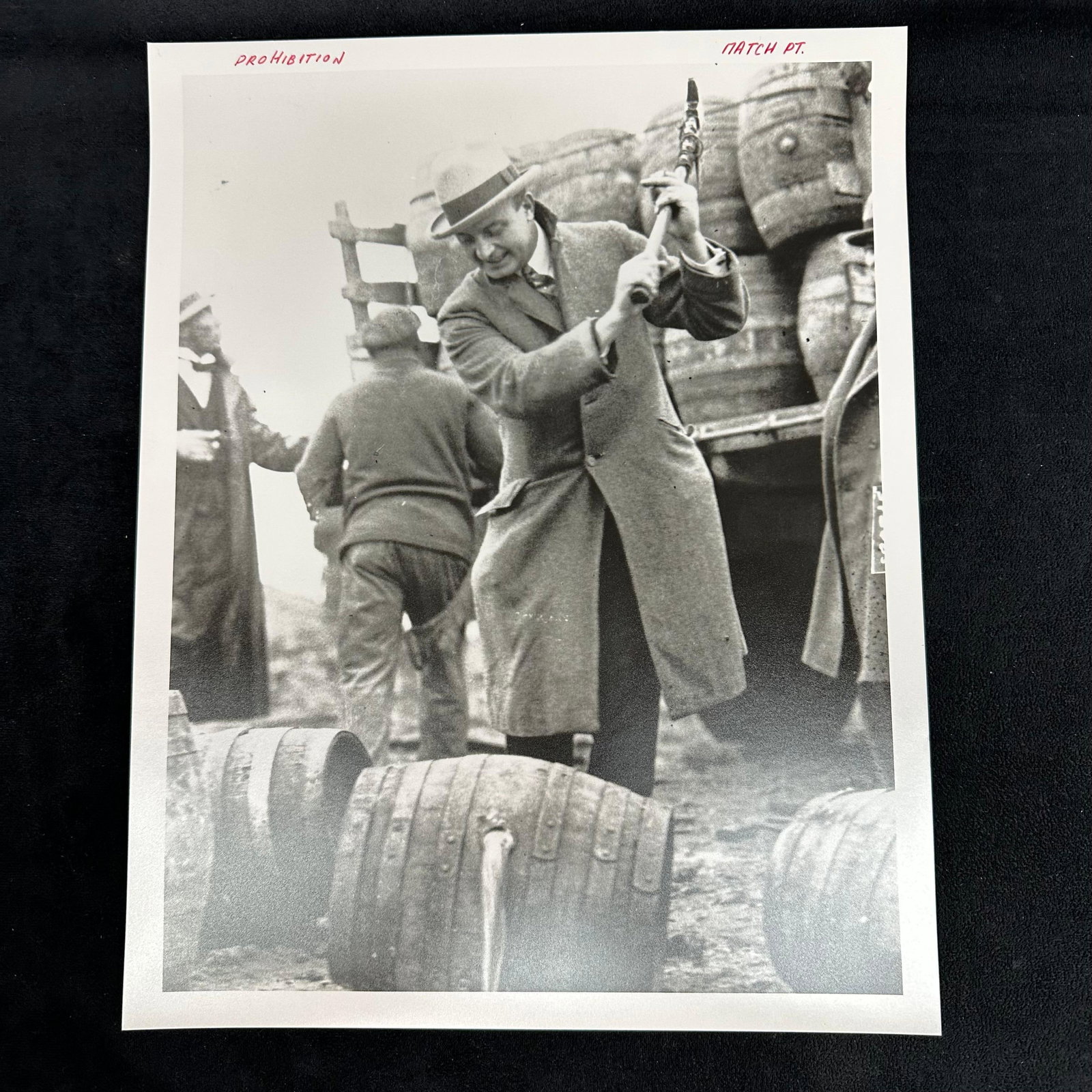 Prohibition Era Photograph Print Officer Smashing Barrels of Alcohol: Black and white analog photo print depicting a Prohibition-era enforcement scene, where a man in a coat and fedora is captured mid-swing while smashing barrels of alcohol with an axe. In the backgroun