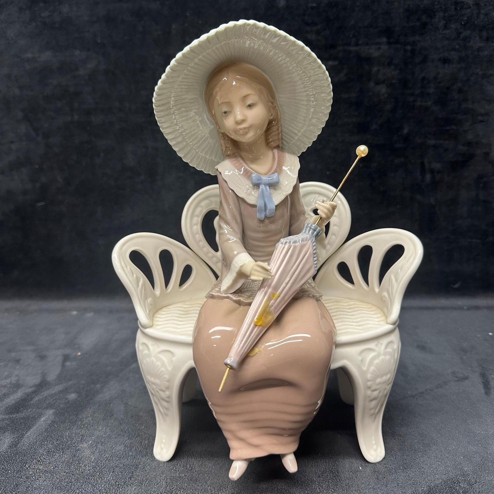 Lladro Waiting in the Park Girl on Bench with Hat and Umbrella: Hallmark of the bottom of the figure. The girl's umbrella is broken, loose piece, with a broken area showing light glue residue. Light markings under the bottom of the figure. This item is in pre-