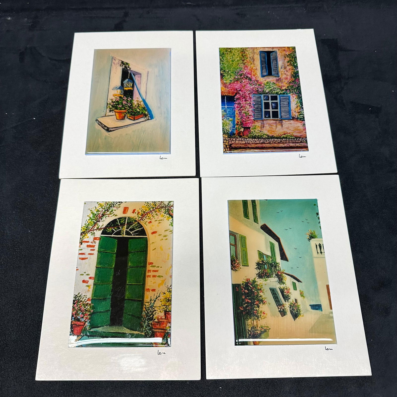 Set of Four Signed European Village Scene Art Prints Lou on Rigid Plastic Acrylic Board: A set of four color art prints depicting European-style village architecture, featuring windows, doors, and street scenes with floral elements. Each print is mounted on a rigid plastic or acrylic boar