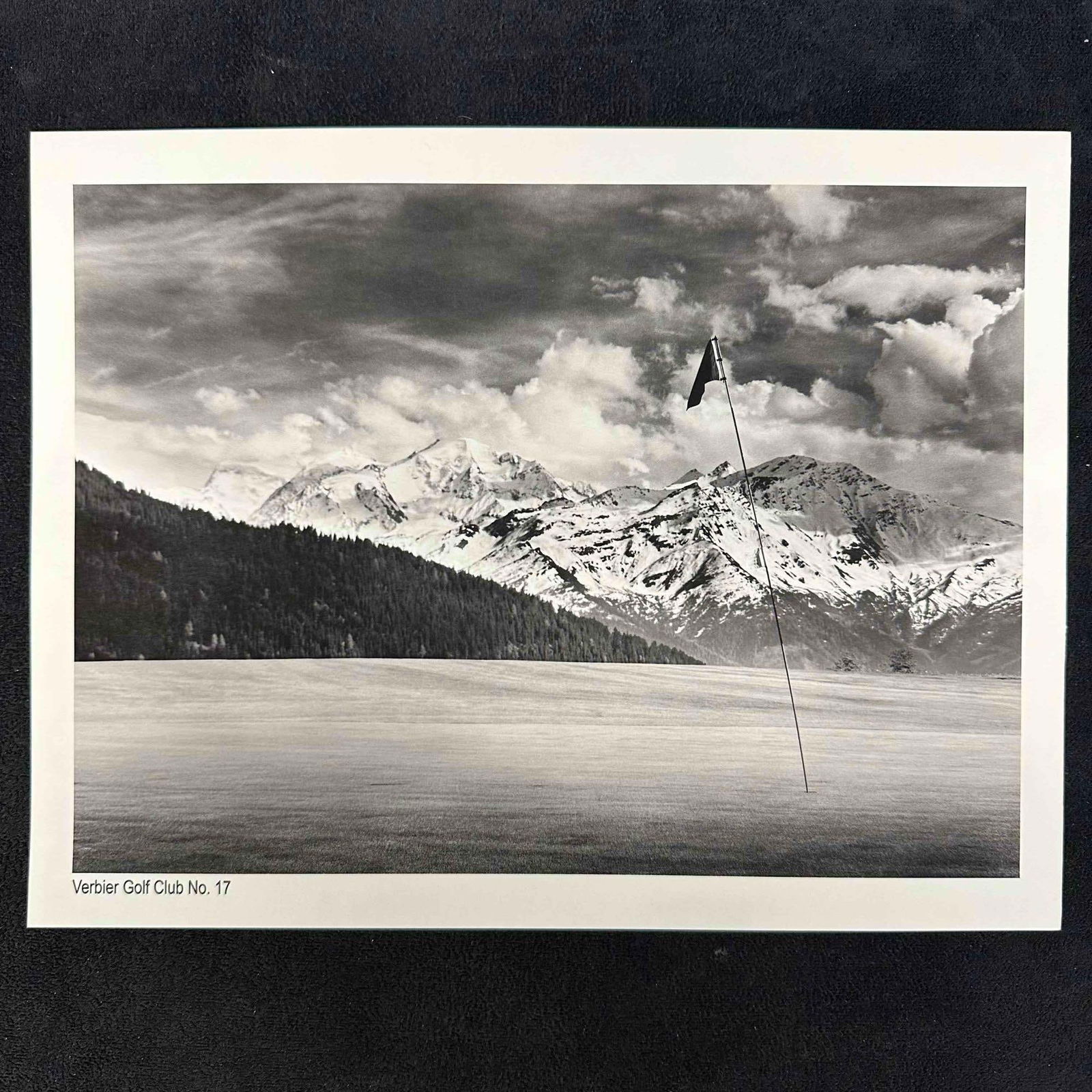Verbier Golf Club No. 17 Black and White Golf Photography Print: Black-and-white photograph featuring the 17th hole at Verbier Golf Club, set against snow-covered peaks and dramatic cloud formations. The lone flag and sweeping fairway create a bold minimalist compo
