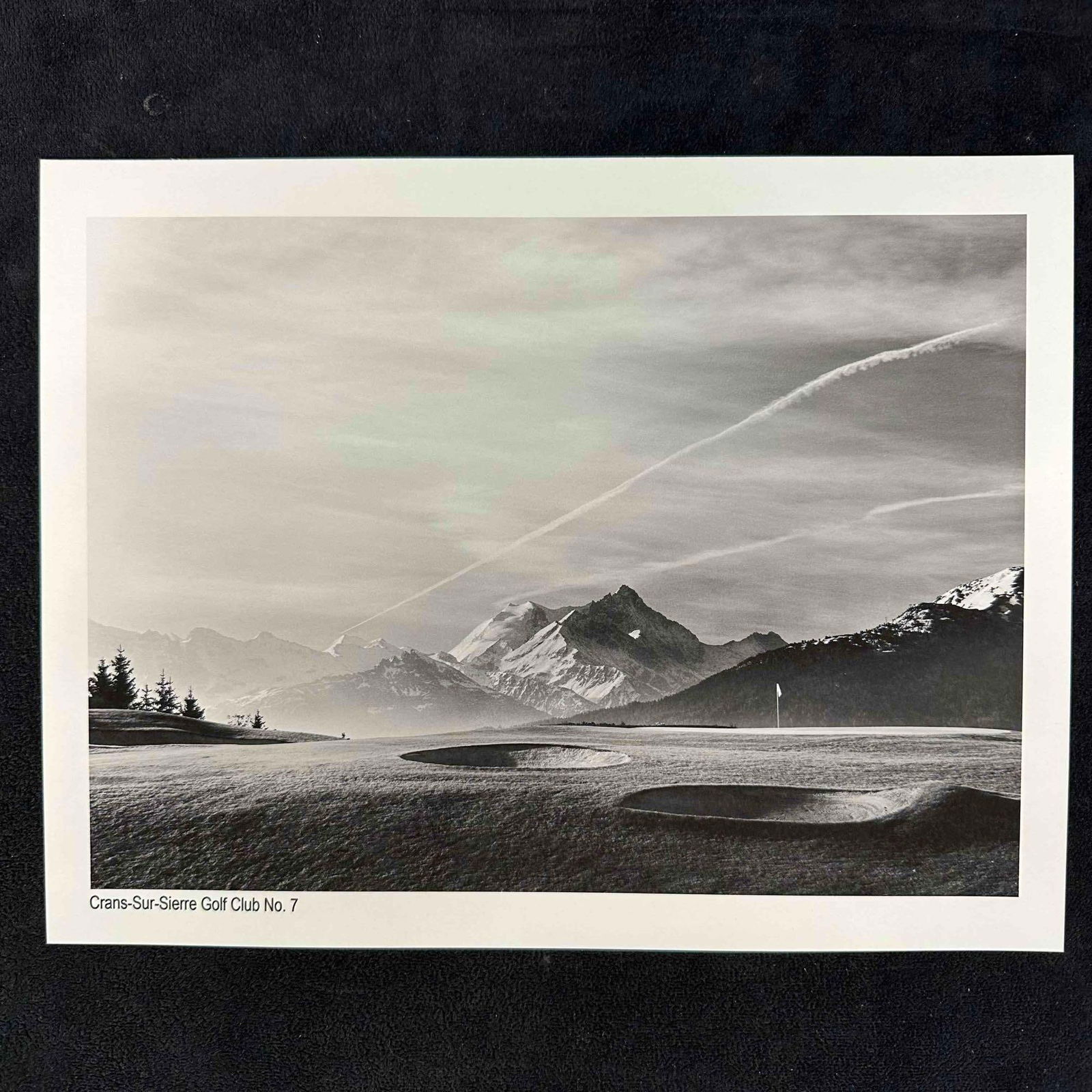 Crans-Sur-Sierre Golf Club No. 7 Black & White Landscape Photography Print: Black-and-white photograph featuring Crans-Sur-Sierre Golf Club’s 7th hole with dramatic mountain views in the background. The image beautifully captures the contrast between the rolling greens, dee