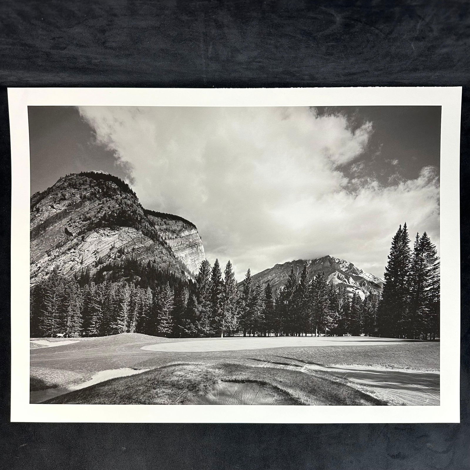 Black and White Inkjet Photograph Print Mountain Golf Course with Sheer Cliff Face: Black and white inkjet print depicting a golf course framed by towering cliffs and evergreen forest. The rugged rock face rises behind the tree line, evoking the grandeur of alpine landscapes. Wide