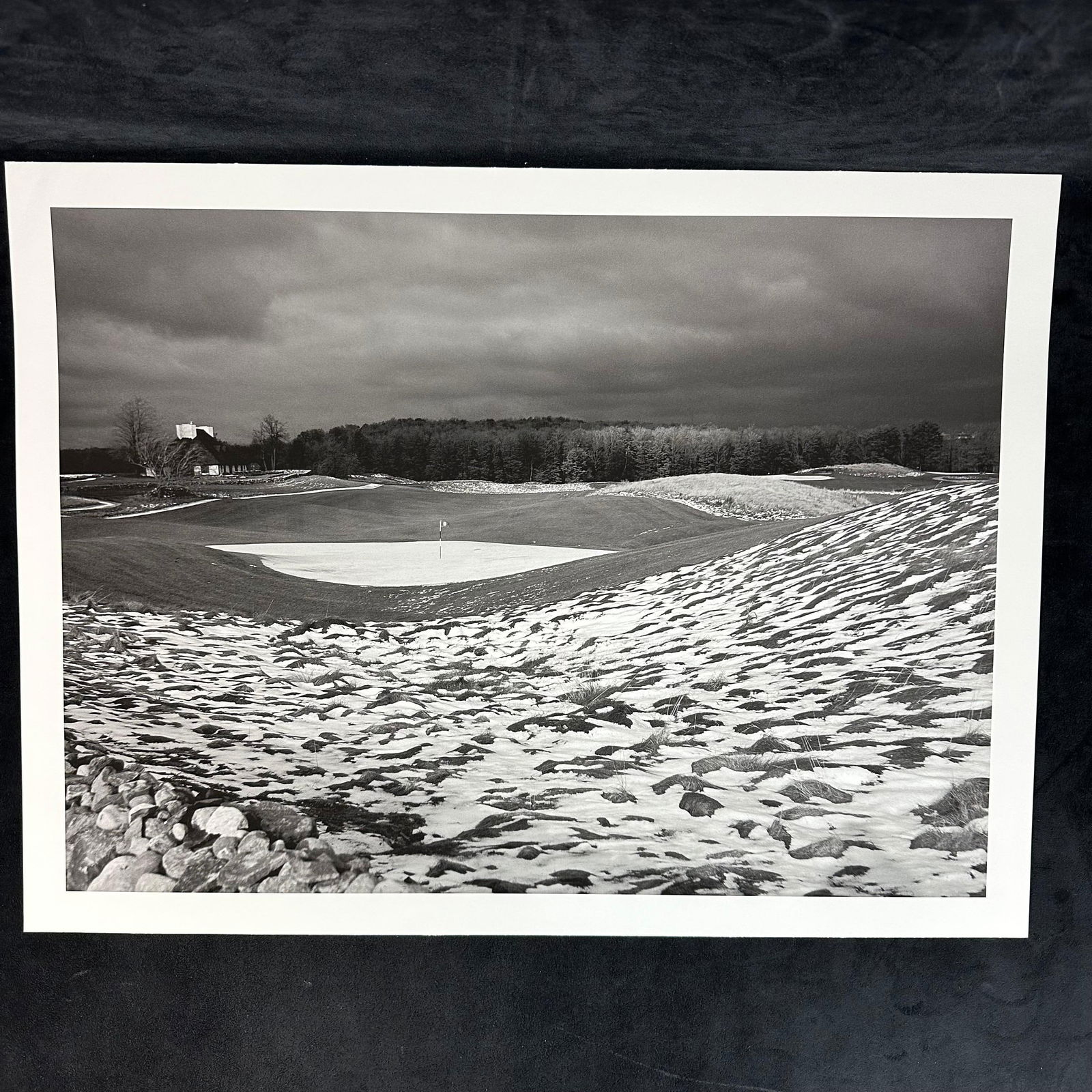 Snow-Dusted Golf Green with Clubhouse in Distance Inkjet Photo Print: Black-and-white inkjet photographic print depicting a striking golf course landscape in winter. The scene shows a green surrounded by rolling hills with patches of snow scattered across the ground. A