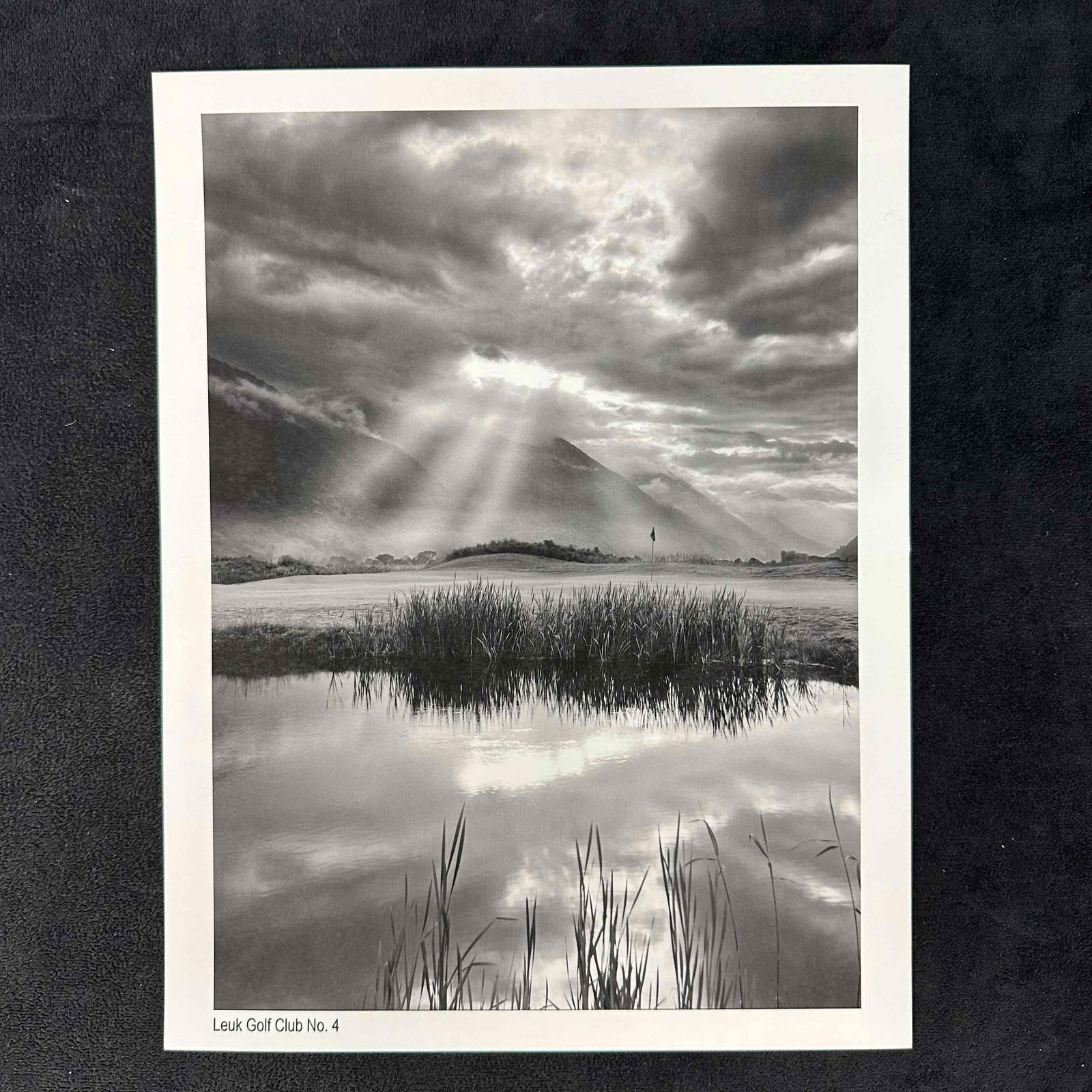 Leuk Golf Club No. 4 Black and White Golf Photography Print: Black-and-white photographic print of Leuk Golf Club’s No. 4 hole, featuring dramatic sun rays piercing through the clouds over a serene mountain backdrop. The foreground shows a reflective pond