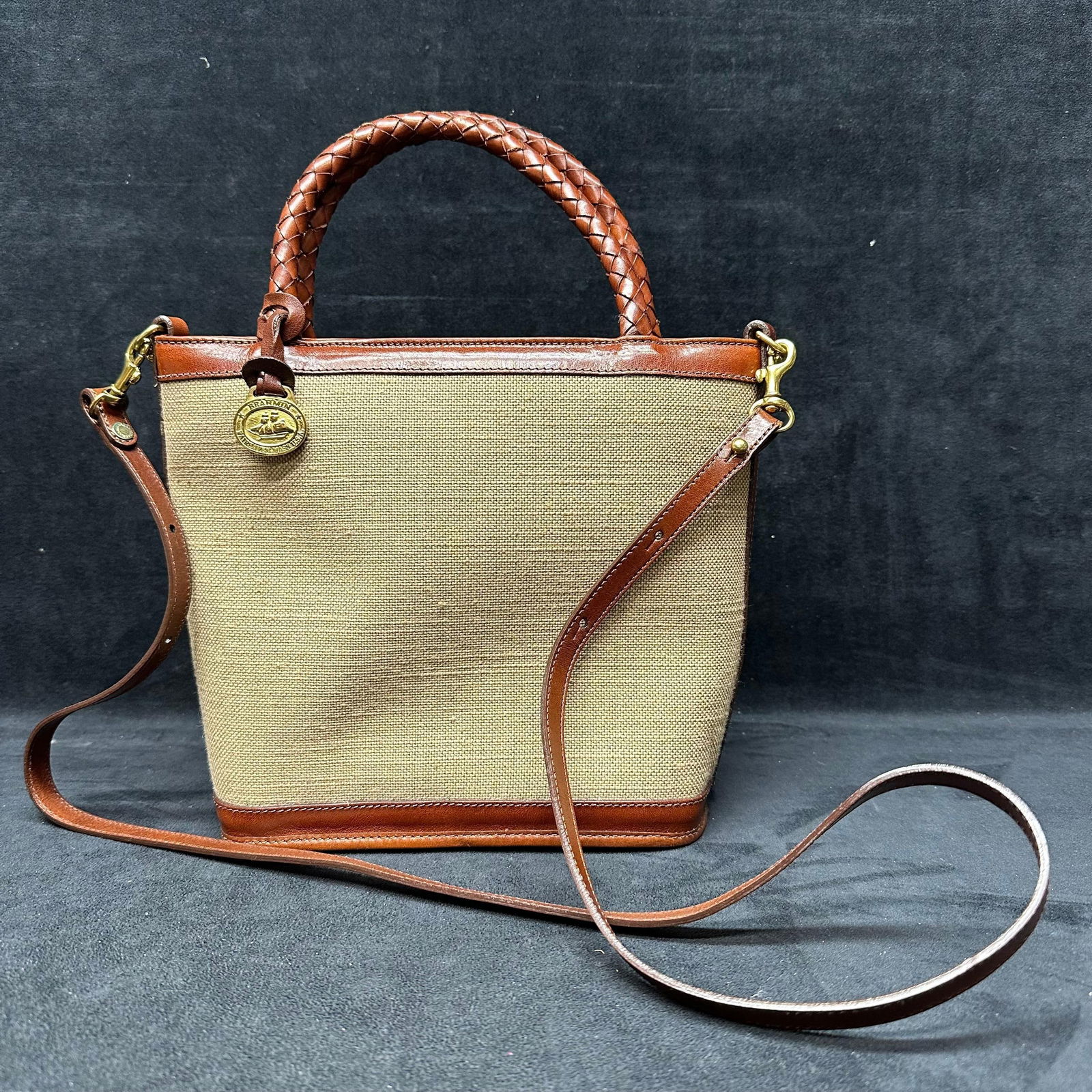 Brahmin Canvas and Leather Tote with Braided Handles and Shoulder Strap: Brahmin structured tote handbag featuring a natural woven canvas body with brown leather trim. Designed with braided leather top handles and a detachable adjustable leather shoulder strap. Accented wi