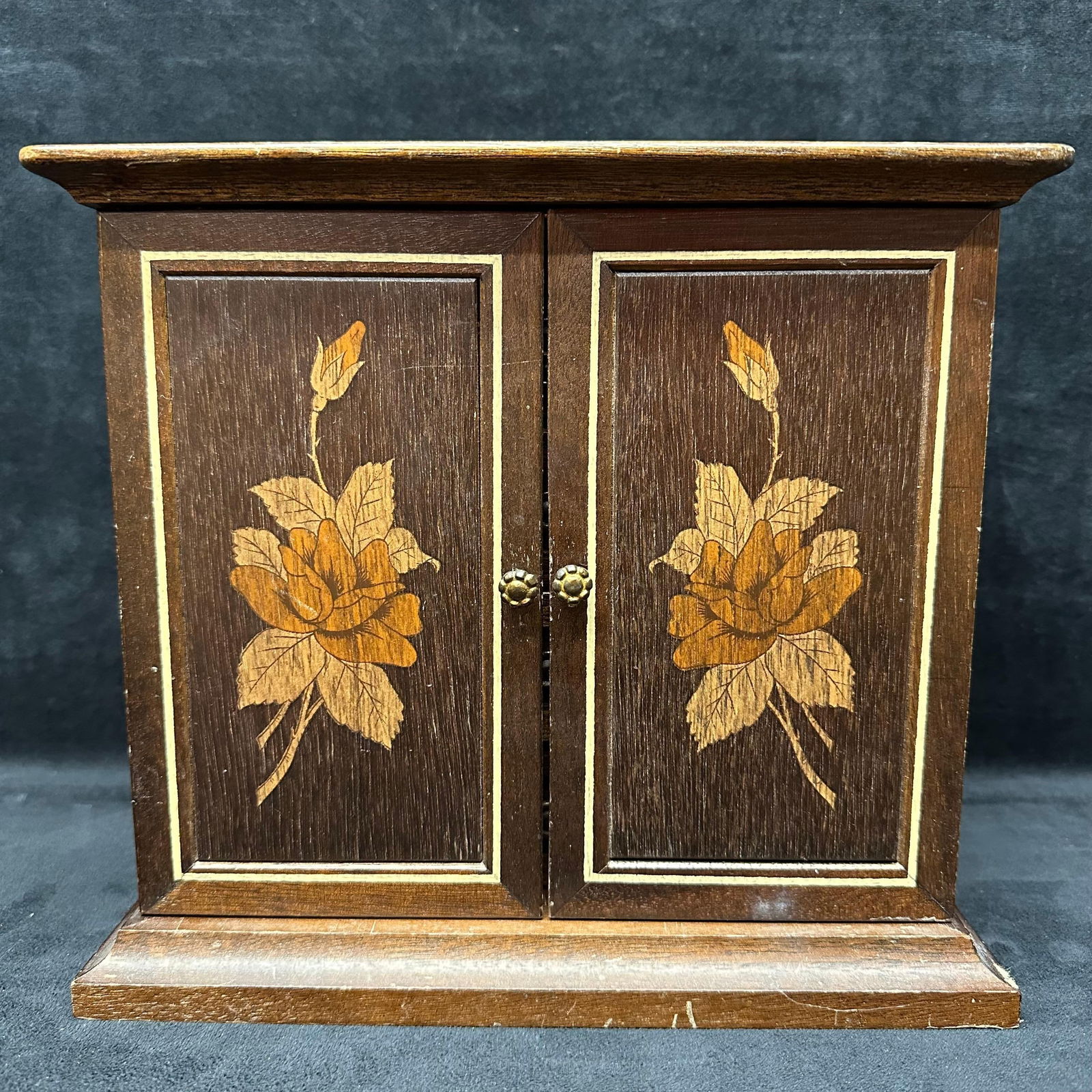 Wooden Jewelry Box with Floral Inlay Design and Red Velvet Lined Drawers: Wooden jewelry box featuring a two-door cabinet with floral inlay details. Inside are four pull-out drawers, each lined in red velvet, for rings, necklaces, or keepsakes. The piece rests on a wood-mol