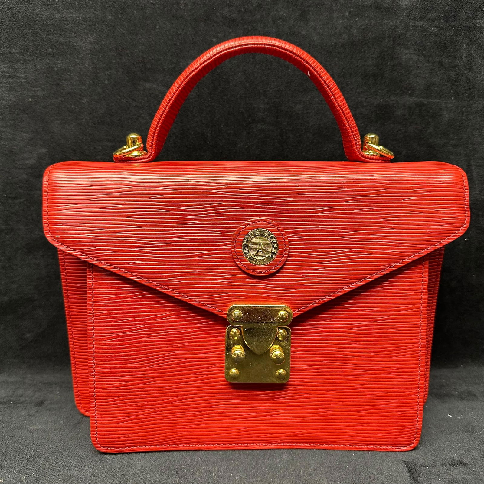 La Tour Eiffel Red Textured Leather Style Top Handle Satchel Handbag: La Tour Eiffel brand structured handbag crafted in red textured leather, with a style mix of gold-tone hardware. The bag features a top handle, a flap-front design with a turn-lock style closure, and