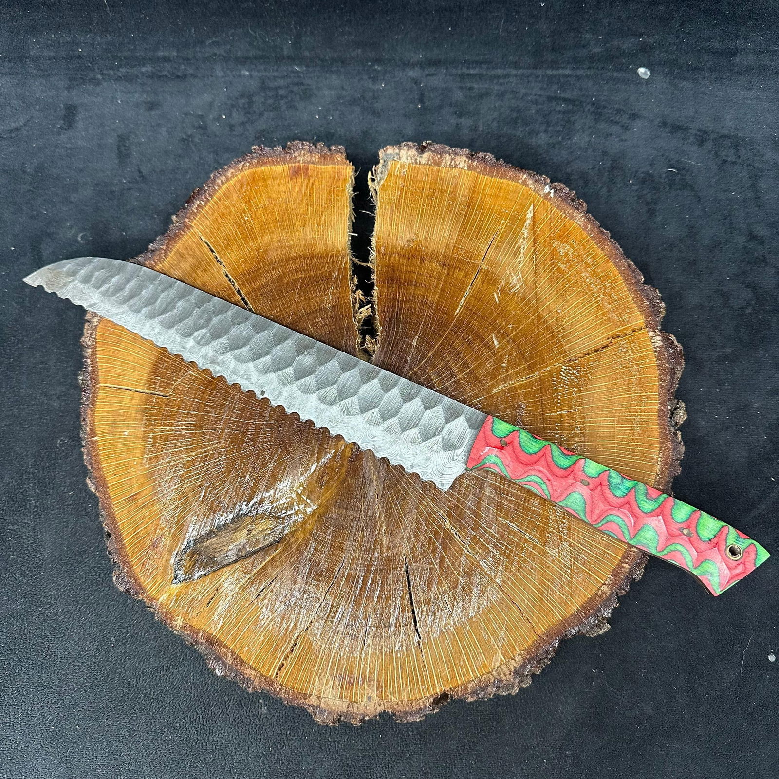 Long Custom Hand-Forged Steel Serrated Knife with Red and Green Patterned Handle: Hand-forged custom knife featuring a patterned steel blade with a hammered finish and serrated spine detail. The blade shows layered steel patterning throughout and a pronounced tapered tip. The