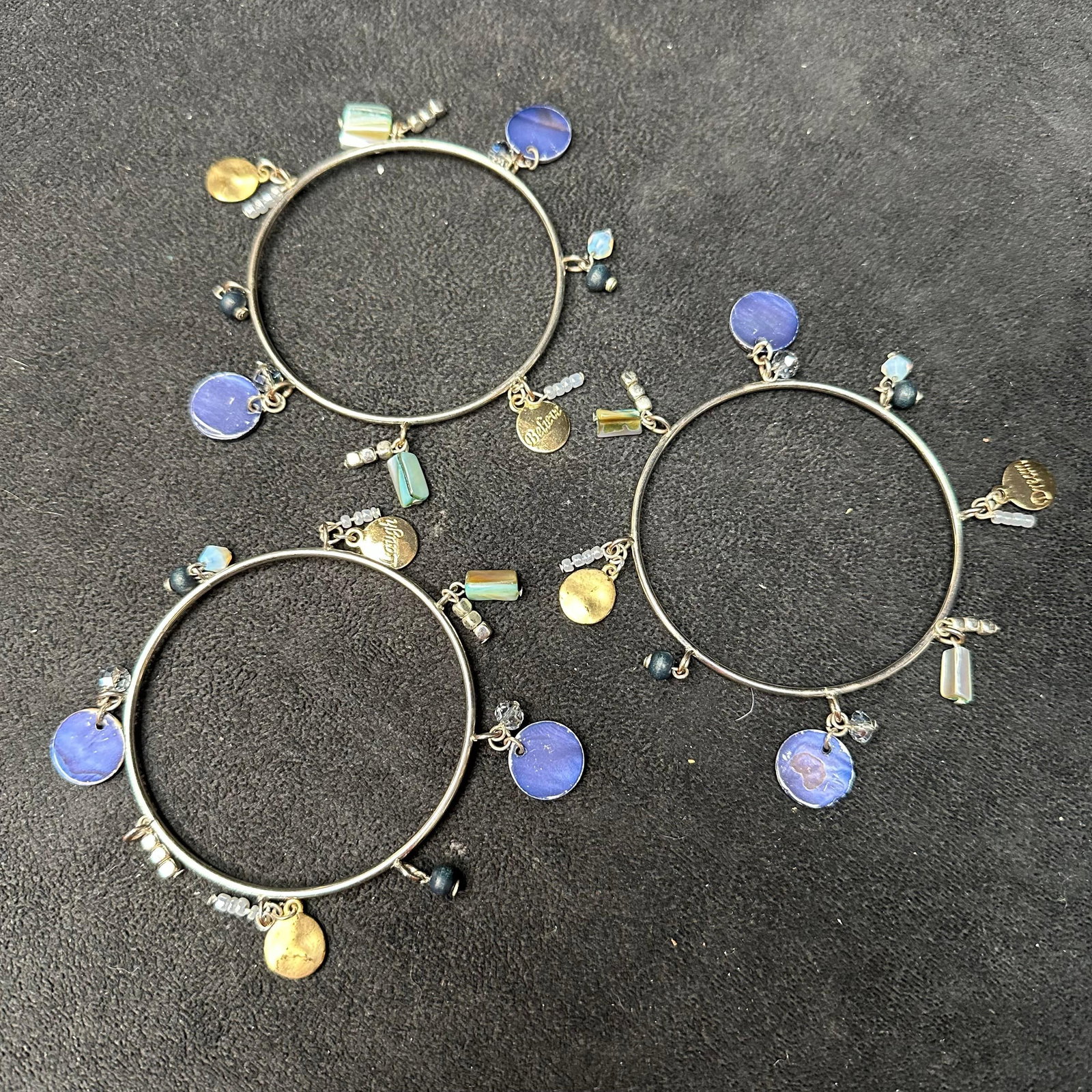 Set of Three Silver Tone Charm Bangle Bracelets with Blue and Gold Dangle Accents: Set of three coordinating silver-tone bangle bracelets featuring assorted dangling charms. Each bracelet is accented with blue enamel or glass discs, faceted beads, small metallic spacers, and