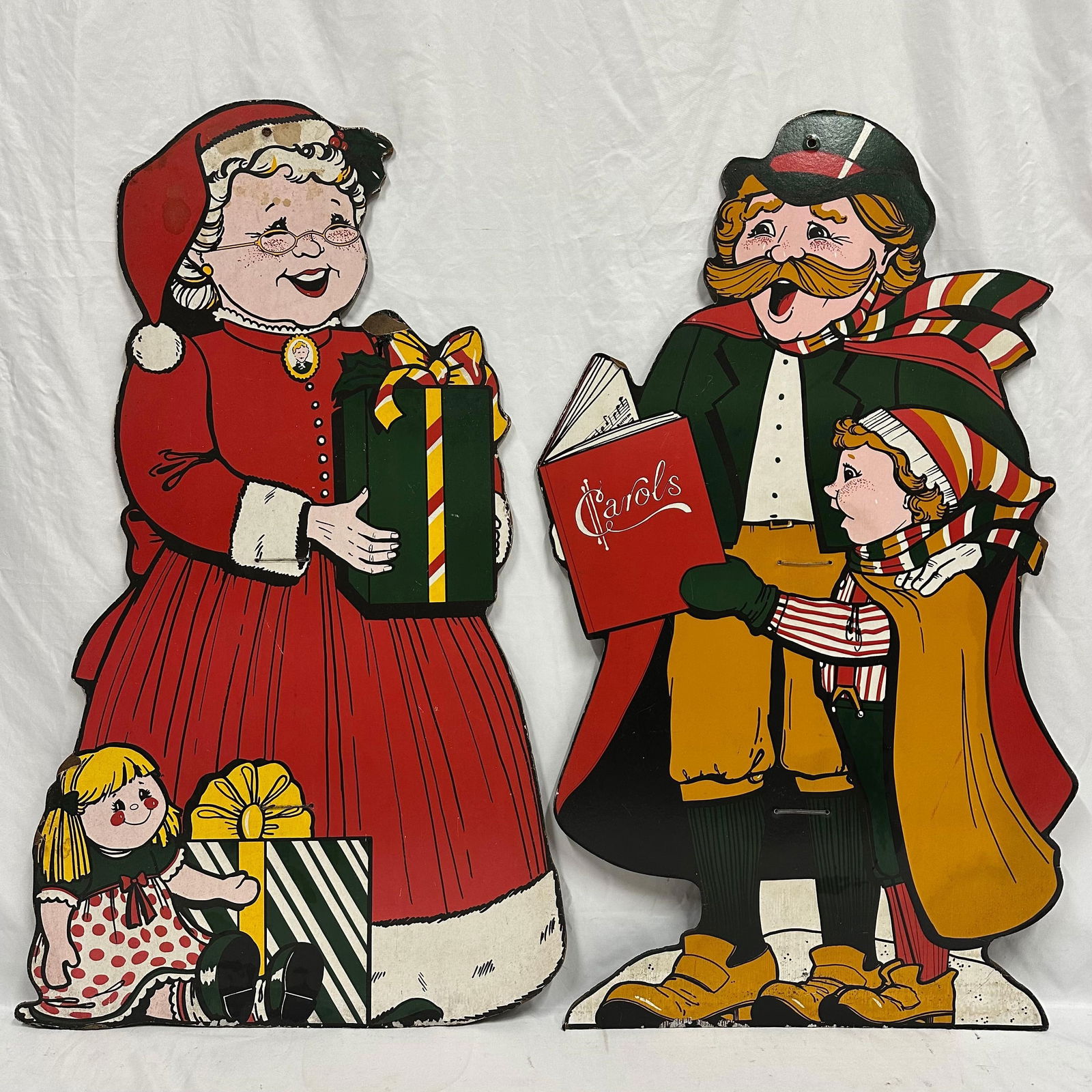 Vintage Christmas Cutout Board Decorations Mrs. Claus and Carolers Set: Pair of vintage Christmas cardboard cutout decorations depicting Mrs. Claus with gifts and a caroling family scene. Vintage holiday decor. Shows age-related wear, surface toning, staining, and edge