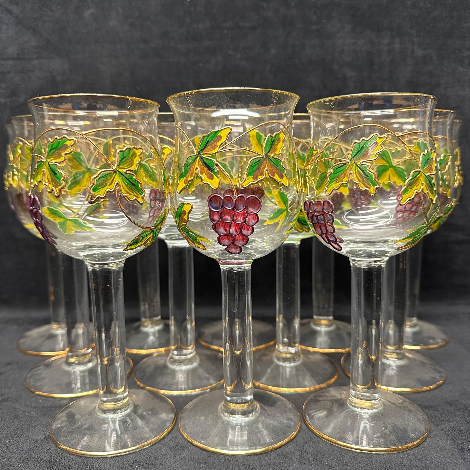 Set of Twelve Hand Painted Grape Motif Wine Glasses with Gold Gilt Accents: Matching set of twelve clear, thick-stemmed wine glasses featuring hand-painted grape clusters and vine leaves in rich green, yellow, and burgundy tones. Each glass is accented with a gold-painted rim