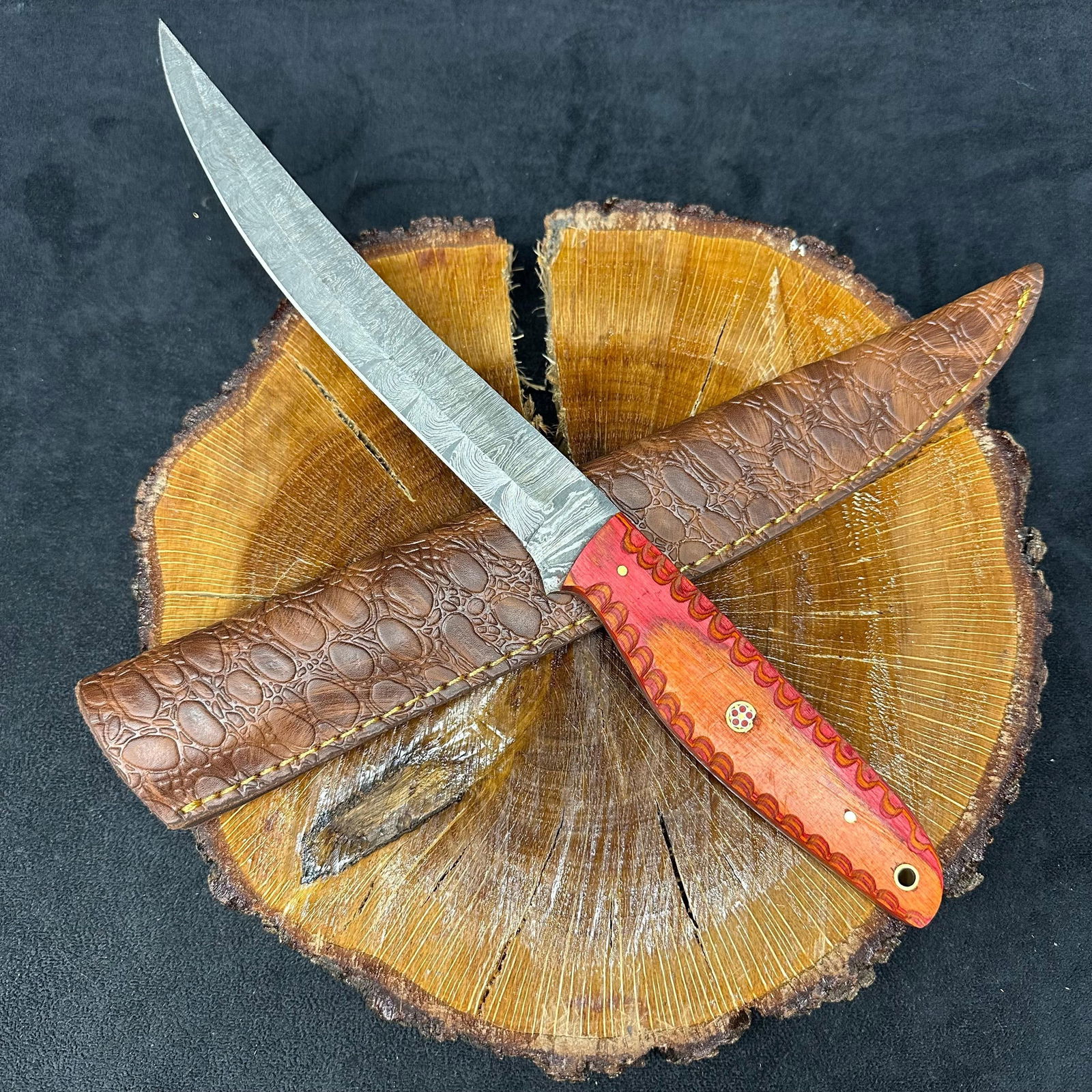 Long Steel Straight Edge Knife with Orange Pattern Handle Q32: A custom knife featuring a patterned steel blade with a hammered finish and a straight spine. The handle is constructed from a contoured, orange-patterned epoxy resin/wood composite material, secured