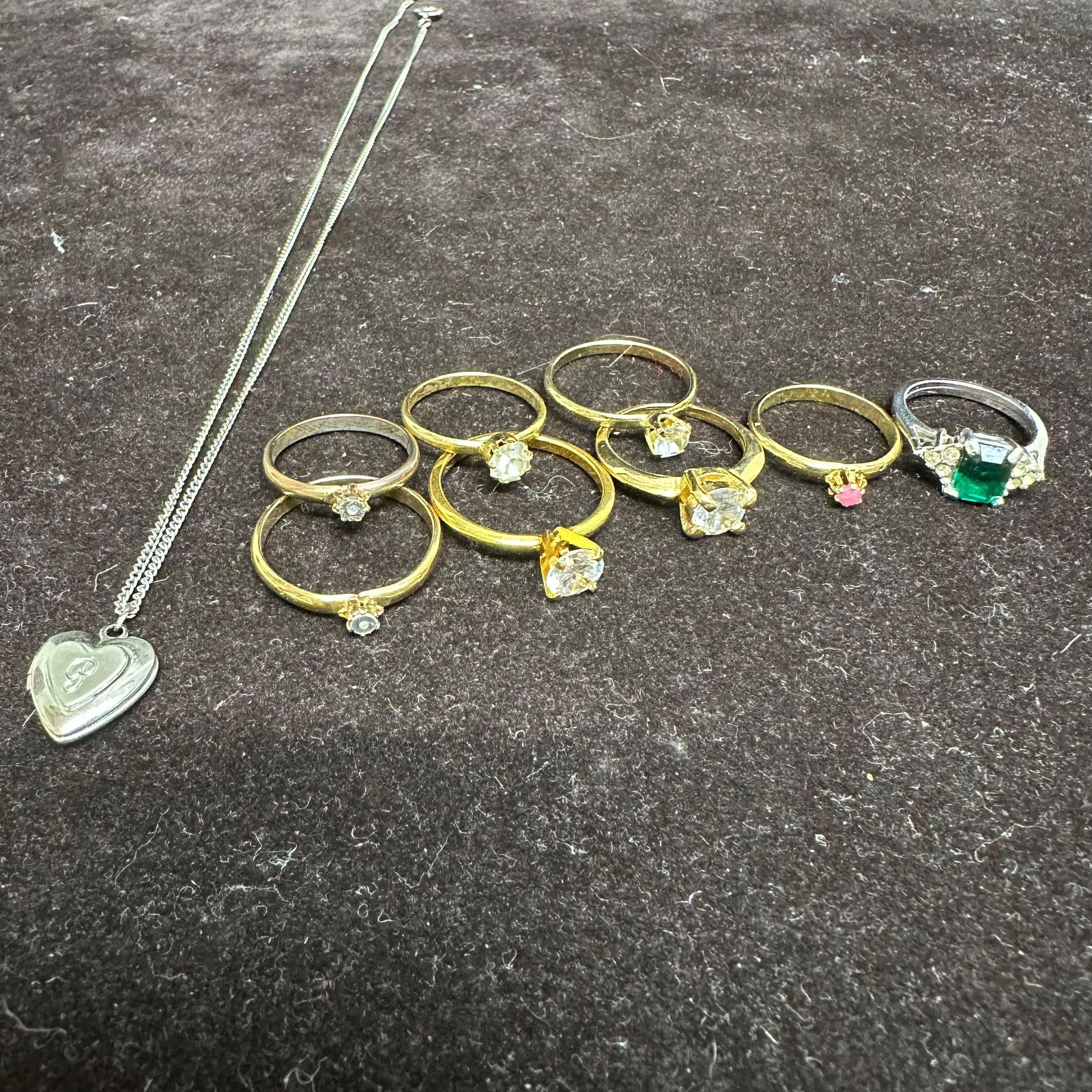 Collection of Costume Jewelry Rings and Silver Toned Heart Locket Necklace: An assorted group of costume jewelry, including seven gold-tone rings and one silver-tone ring set with simulated gemstones, featuring clear, pink, and green stones in various cuts. Also included is a