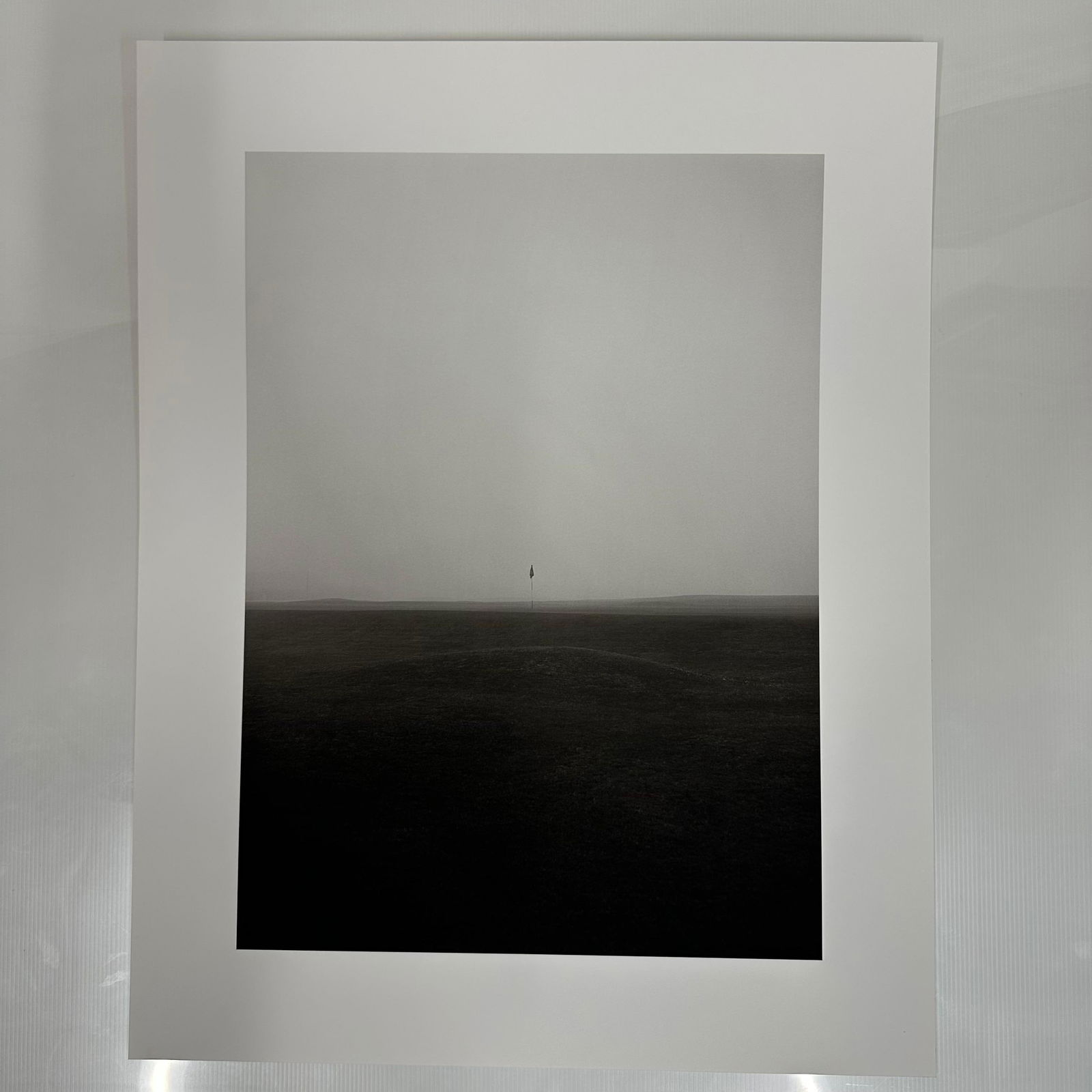 Black and White Minimalist Golf Course Photography Print Foggy Fairway Scene #3: Black and white photographic print depicting a solitary golf flag standing on a mist-covered fairway. The minimalist composition and muted tones create a serene, almost abstract landscape. Printed on