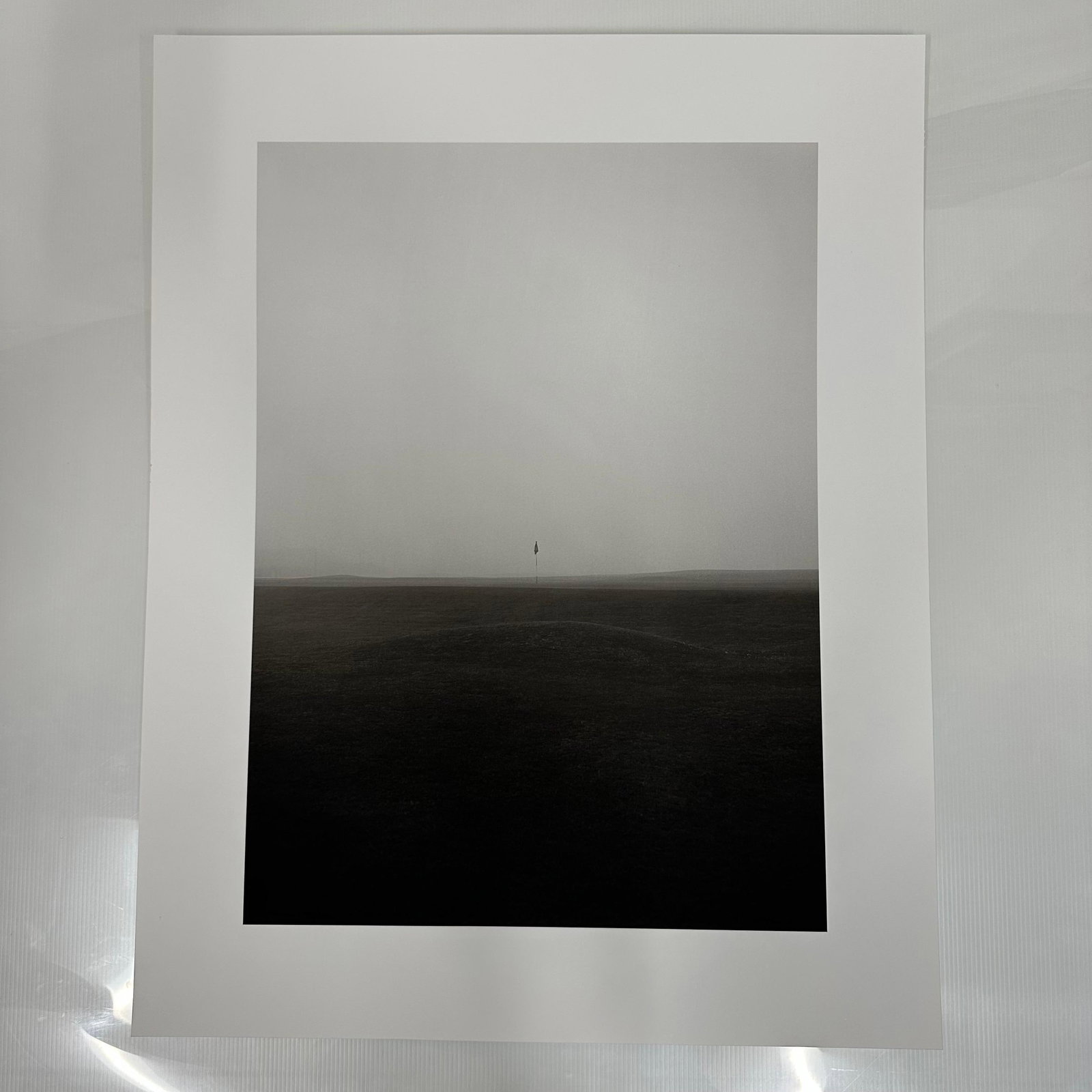 Foggy Golf Course Fairway Scene Minimalist Black and White Photography Print #2: Black and white photographic print depicting a solitary golf flag standing on a mist-covered fairway. The minimalist composition and muted tones create a serene, almost abstract landscape. Printed on