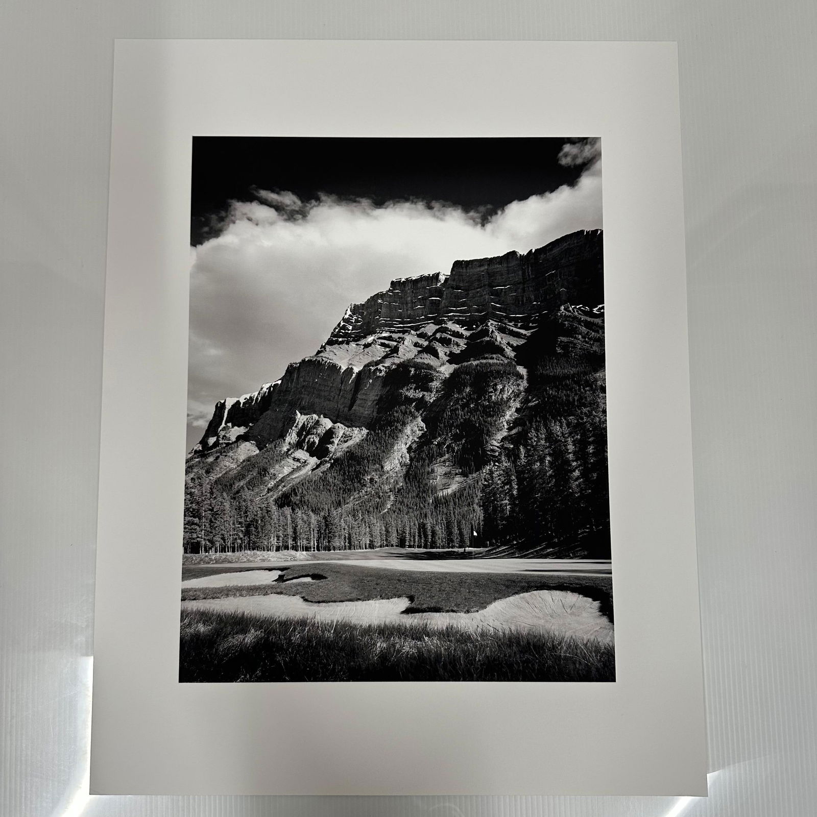 Black and White Landscape Photography Print Mountain Golf Course Scene: Black and white photographic print depicting a dramatic mountain range rising above a golf course landscape, possibly the Banff Springs Golf Course in Alberta, Canada. The image captures the contrast