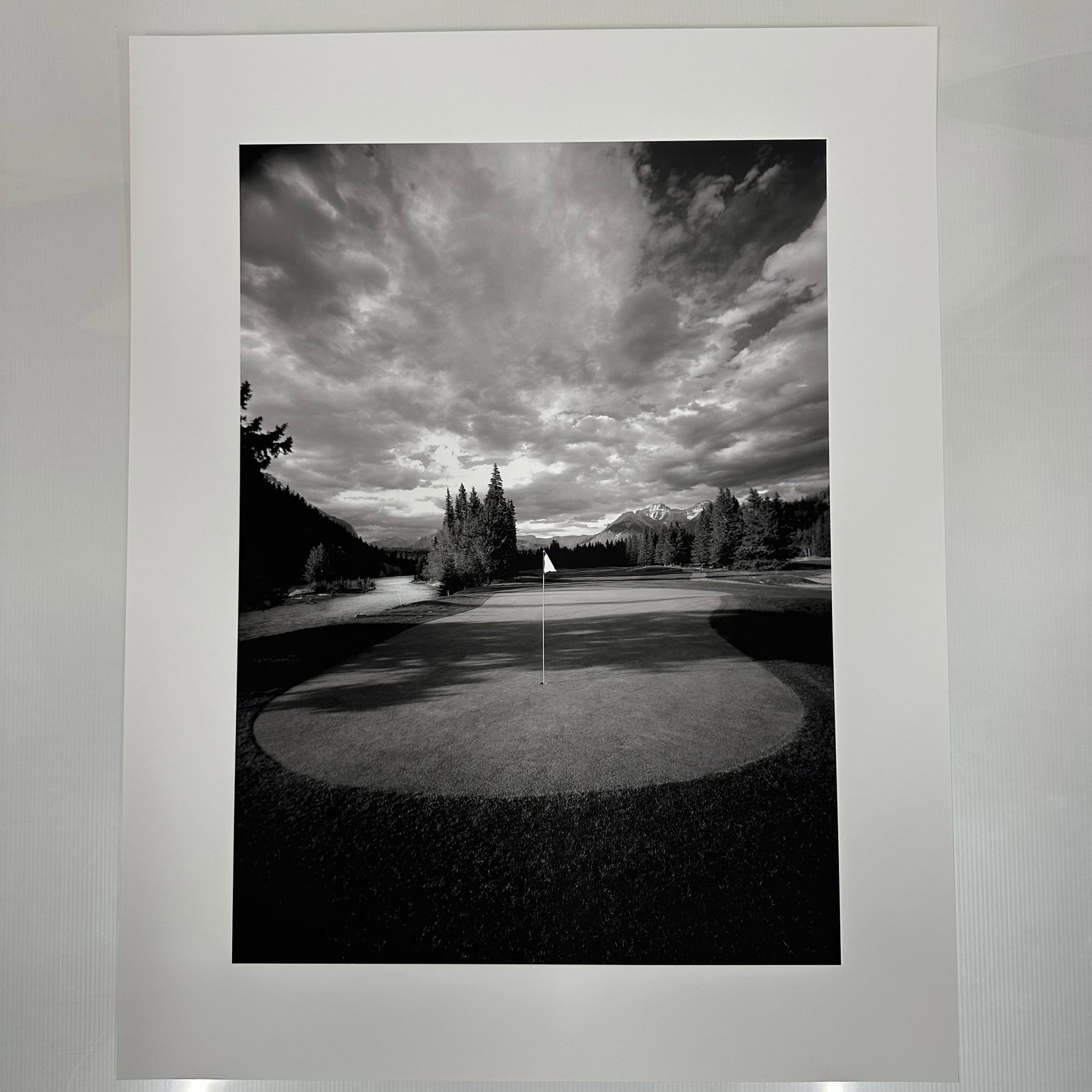 Banff Springs Golf Course in the Rocky Mountains of Canada Black and White Photo Print A: Large format black and white fine art photography print showing a golf green with a centered flag, surrounded by trees and distant mountains under a dramatic clouded sky. Printed on matte photo paper