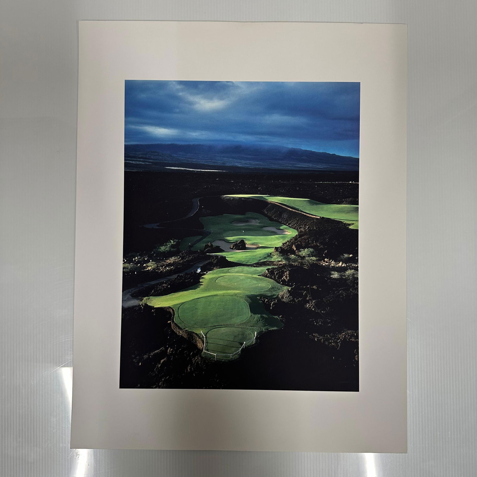 Aerial Golf Course Landscape Over Fairways Fine Art Color Photography Print: Vivid color photograph depicting an aerial view of a golf course set against dark rocky terrain and a dramatic twilight sky. The striking contrast of emerald greens and deep blues captures the serene