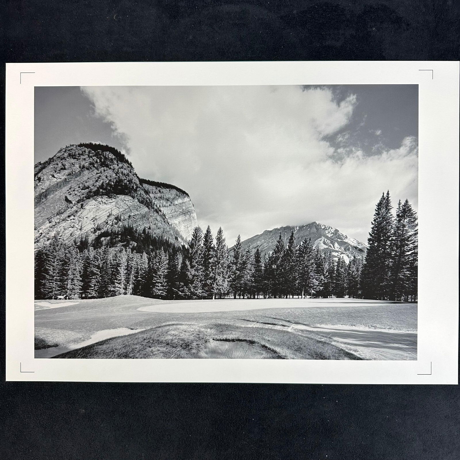 Mountain Golf Course Landscape Black and White Photography Print: Black and white landscape photography print capturing a golf course framed by towering mountains and dense evergreen forests under a partially clouded sky. Printed on high-quality photo paper, blank r