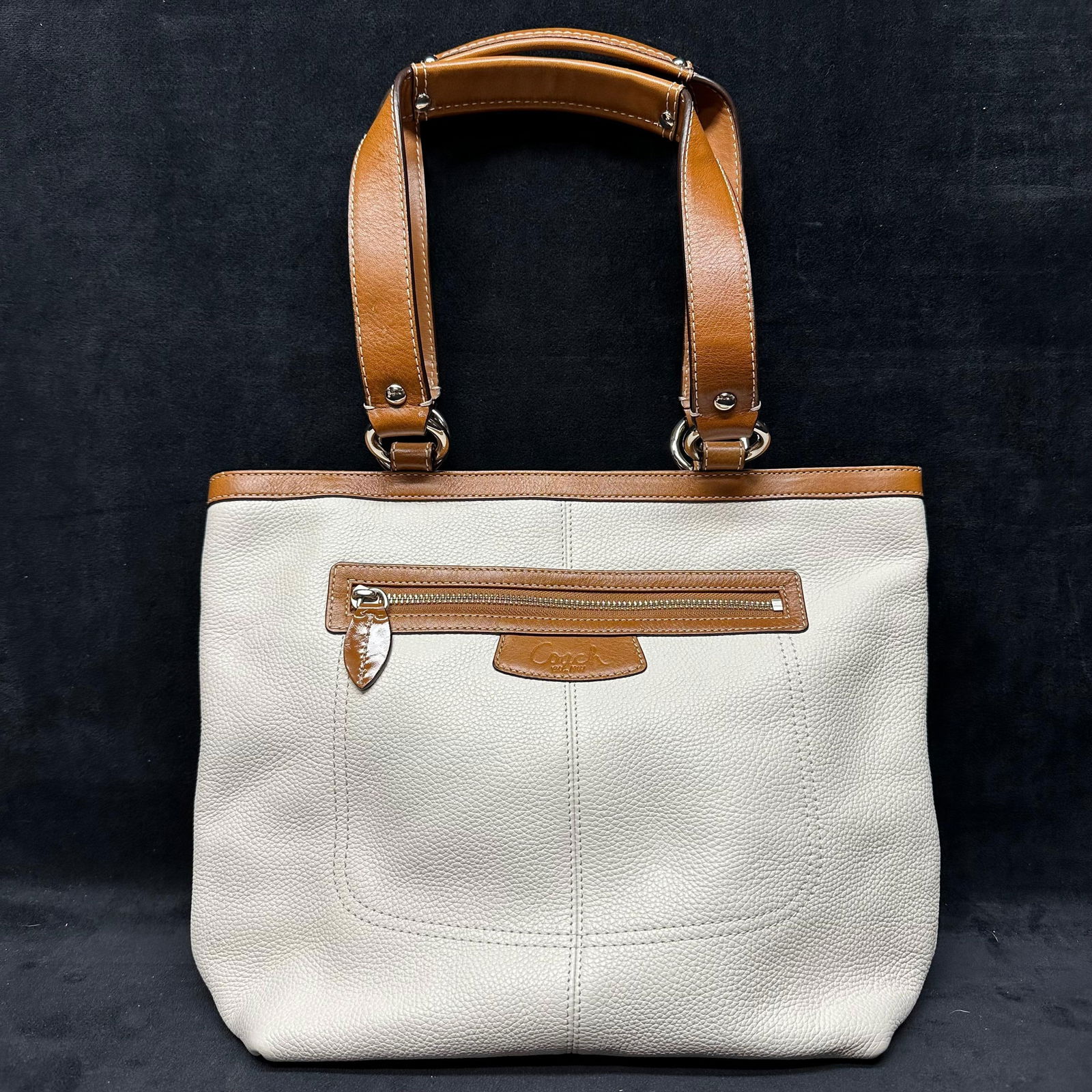 Coach Two-Tone Pebbled Leather Tote Bag Cream and Tan Colored: Coach tote bag crafted in pebbled leather with a cream-colored body and contrasting tan leather trim and handles. Features double shoulder handles with silver-tone ring hardware, a top zip closure, an