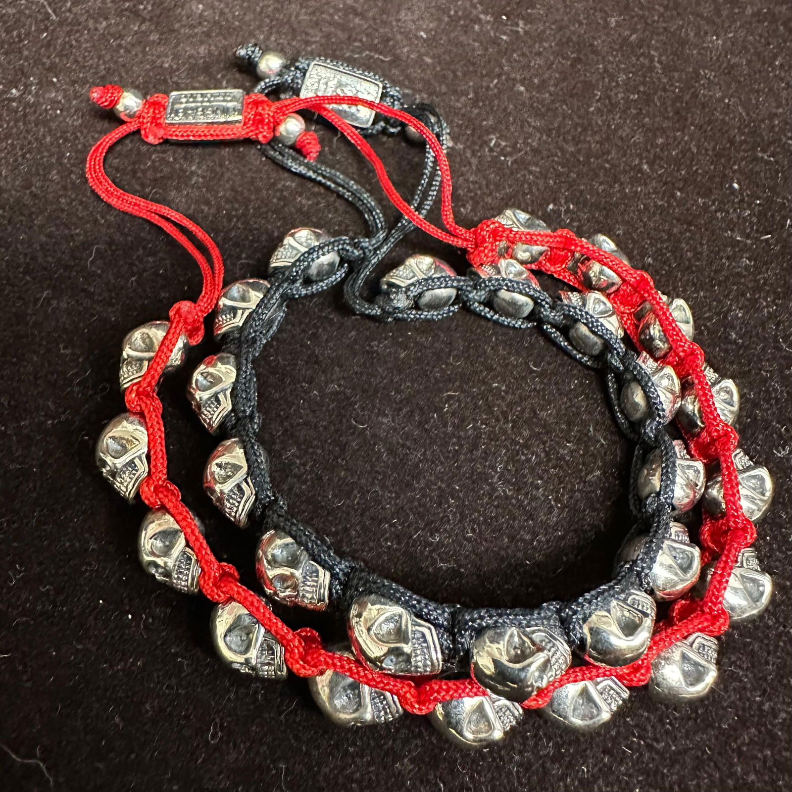 King Baby Studio Silver Tone Skull Red Black Macrame Braiding Bracelets: Pair of King Baby Studio skull bracelets featuring multiple cast skull beads strung on braided cord. One bracelet is red cord, and the other is black cord. Each bracelet has an adjustable sliding clos
